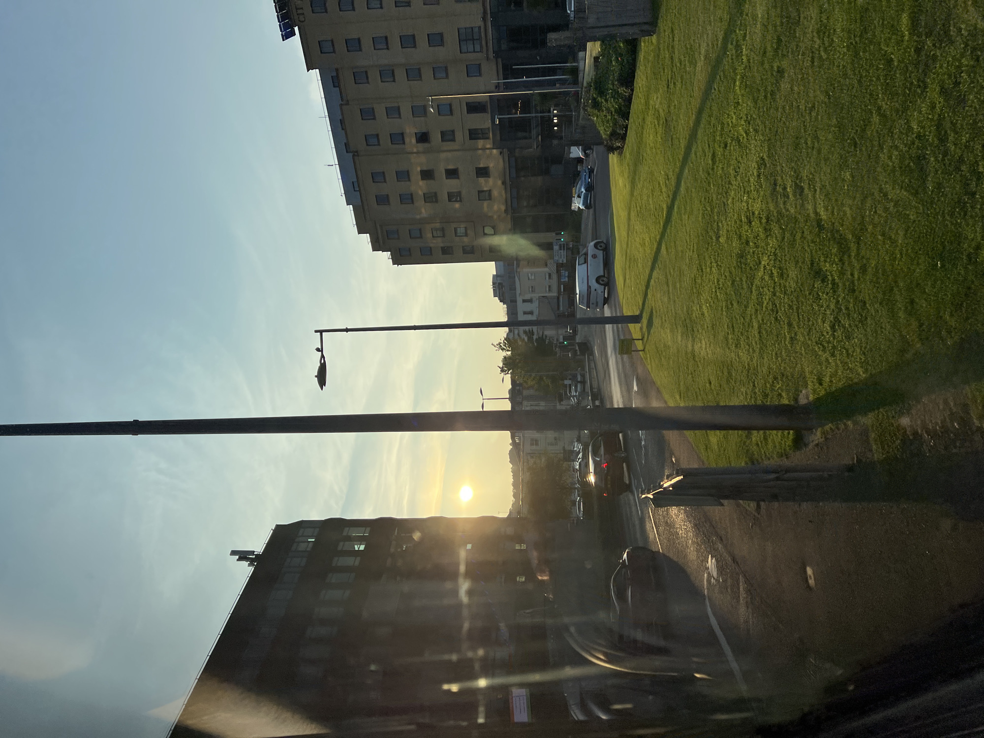 The image captures a street scene in the city of Derry/Londonderry, United Kingdom, during what appears to be sunset. A large, dark building, possibly an office block, dominates the left side of the frame, with the setting sun reflecting off its windows. Across the street, a light-colored, multi-story building, likely a hotel, stands prominently. The street is moderately busy with cars, including a white van, a dark car, and a blue car visible. A well-maintained grassy area is situated in the foreground on the right, with a tall lamppost casting a shadow. The sky is clear with a soft gradient of blue and yellow hues indicating the setting sun. The overall impression is of an urban environment bathed in the warm light of dusk.