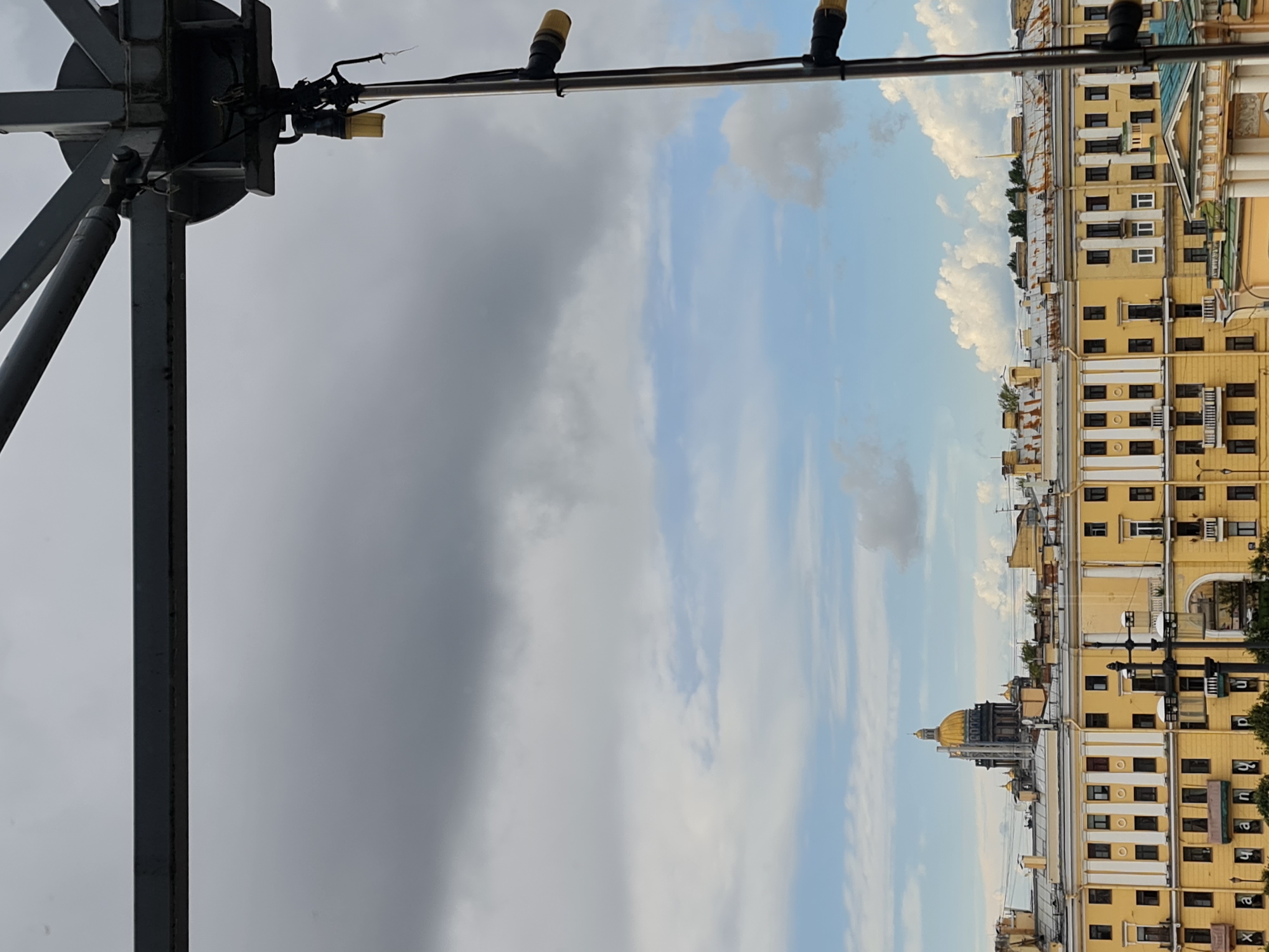 The image captures a view of Saint Petersburg, Russia, likely from an elevated vantage point. Dominating the upper portion of the frame is a large, gray metal structure, possibly part of a scaffolding or a temporary support system. Below this structure, the sky is a mix of dark, heavy clouds and patches of lighter blue sky with scattered white clouds.
In the middle ground, a row of yellow buildings stretches across the scene. These buildings appear to be multi-story with numerous windows and decorative architectural elements like columns and balconies. The roofs are a mix of lighter colored materials and some show signs of rust.
Further in the background, a prominent golden dome of a large building, likely a church or cathedral, is visible above the yellow buildings. This dome, along with the overall architectural style, strongly suggests the location is Saint Petersburg. The scene is devoid of people, and no specific actions or interactions are depicted. The environment suggests an urban setting with a cloudy sky, indicating a potentially overcast day.
