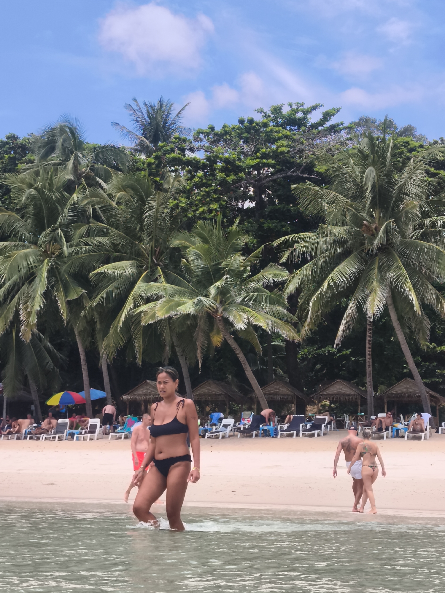 In the city of Ko Samui District, Thailand, a tropical beach scene unfolds under a partly cloudy blue sky. In the foreground, the clear, shallow water of the Gulf of Thailand laps at the sandy shore. A woman in a black bikini is walking out of the water towards the beach. Behind her, a man in red shorts and a woman in a green bikini are walking away from the camera. Further back, a crowd of people are relaxing on lounge chairs under thatched umbrellas, with palm trees lining the edge of the beach. The overall atmosphere is one of a relaxed holiday destination.