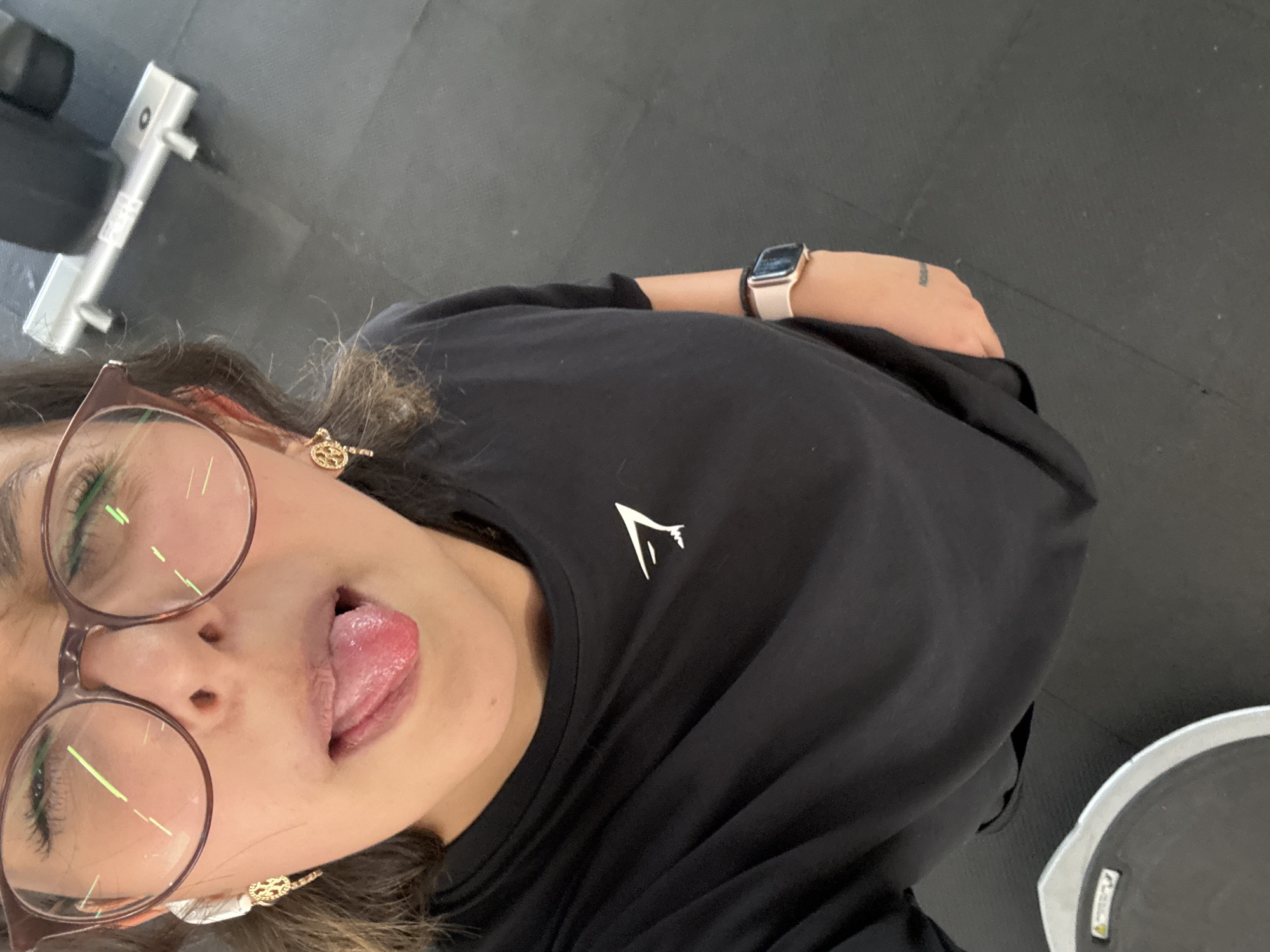 A close-up, high-angle selfie captures a young woman playfully sticking out her tongue, her face angled upwards towards the camera. She has dark hair and wears brown-rimmed glasses, through which green reflections from overhead lights are visible. Her expression is casual and lighthearted. She is wearing a black short-sleeved t-shirt featuring a white "Gymshark" logo on the chest. On her left wrist, she has a light-colored smartwatch with a dark band, and a small, indecipherable tattoo is visible on her forearm. Gold-colored dangling earrings are visible on her ears. The background suggests an indoor gym environment in San Andrés Cholula, Mexico, with a dark floor. A silver piece of gym equipment, possibly a dumbbell or barbell handle, is partially visible in the upper left, and a circular object, potentially a scale or foot pad, with some faint text, is seen in the lower right.