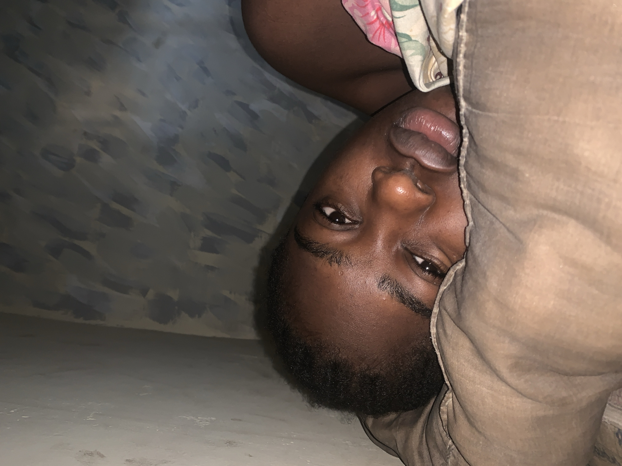 A dark-skinned person with short black hair is lying down, partially covered by a tan-colored blanket or sheet. The person's face is visible, and they appear to be resting their head on the blanket. The background consists of a wall with a textured, painted surface in gray and white tones, and a light-colored floor. The scene is indoors. It's impossible to verify the exact location in Rafin Par, Nigeria without additional location markers or information.