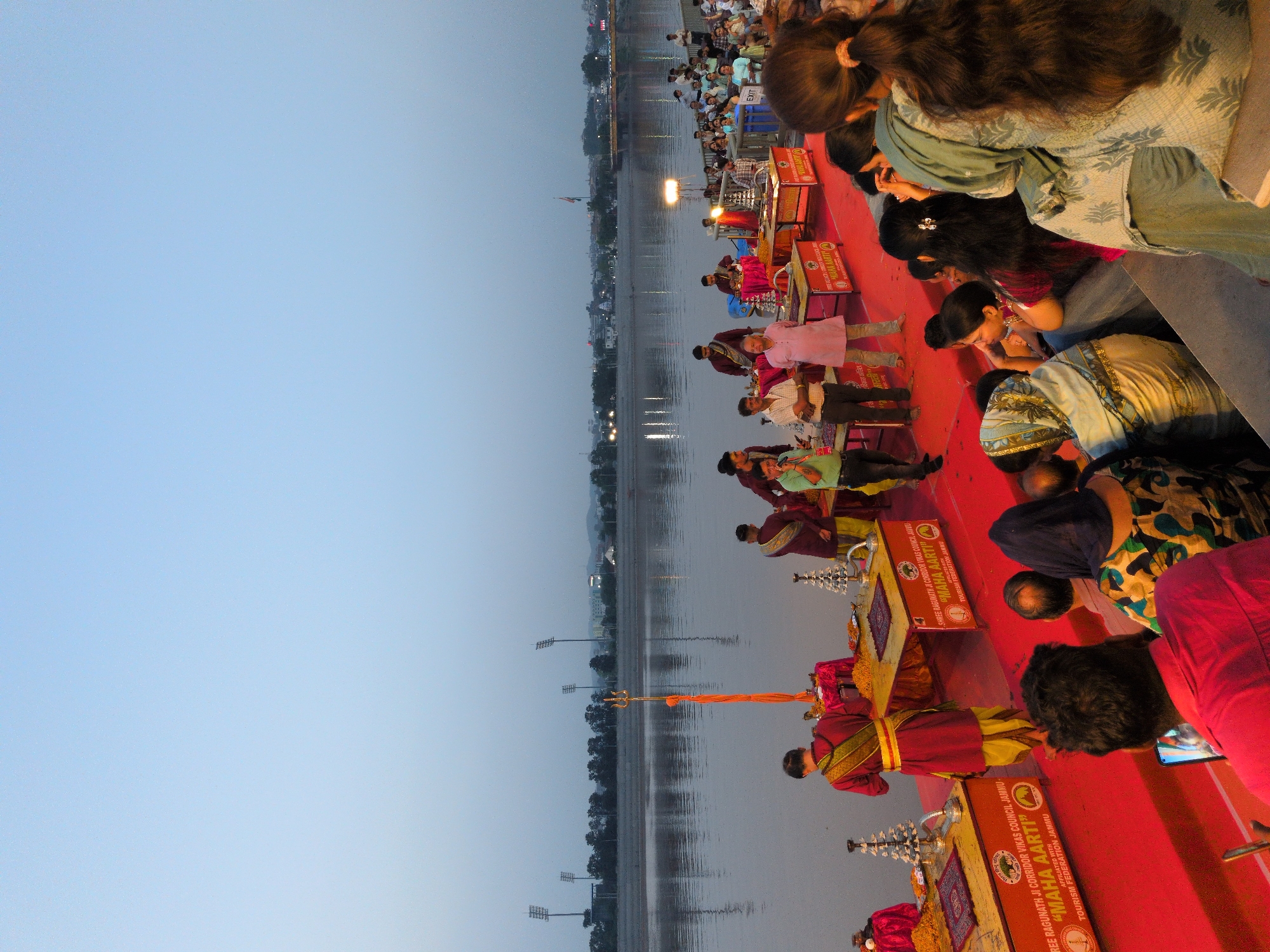 The image depicts a Ganga Aarti ceremony taking place on the banks of the Tawi River in Jammu, India, possibly at the Mubarak Mandi complex. In the foreground, a group of people are seated on red carpeting, with some individuals standing and participating in or observing the ceremony. One man in a pink tunic and another in a red and yellow robe are prominent. Several tables with religious items and offerings are set up. In the background, the Tawi River stretches out, with a cityscape visible on the opposite bank, including lights and a bridge. Tall floodlights and trees line the distant riverbank, and the sky is a pale blue, suggesting either dawn or dusk. The ceremony appears to be well-attended, with a crowd gathered to witness the event.