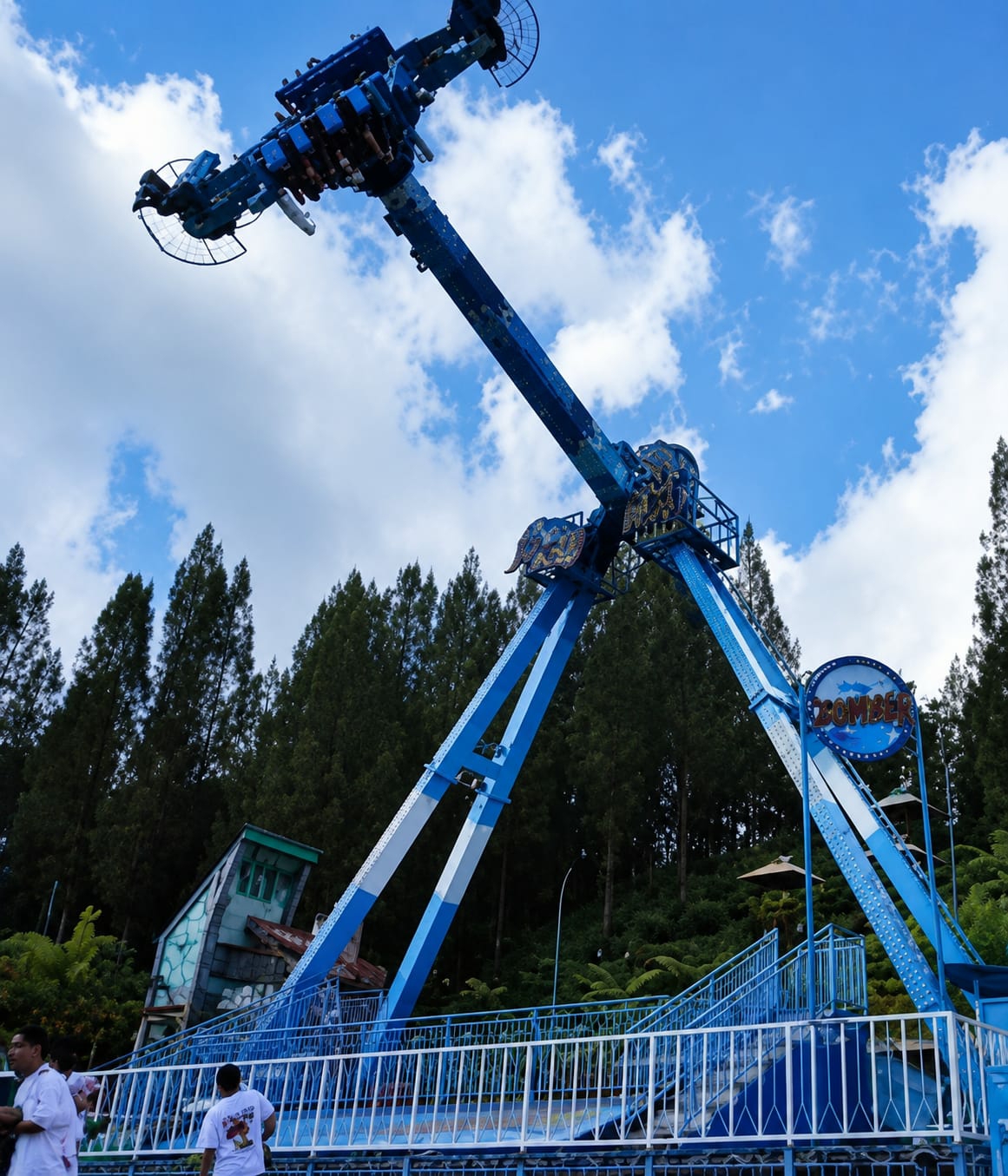 The image shows a large blue amusement park ride, likely a pendulum ride or a similar thrill ride, in operation against a backdrop of a clear blue sky with scattered clouds and a dense forest of evergreen trees. The ride is the central focus, with its arm extended upwards and a gondola of passengers visible at its highest point. The ride structure is predominantly blue with white accents. Below the ride, a white metal railing encloses a waiting area or queue. In the foreground, two men are visible, one wearing a white shirt and the other in a white t-shirt. Behind the railing, a small, whimsical-looking structure with a green and blue facade is partially visible. The environment appears to be an outdoor recreational area, possibly a theme park or an amusement park, situated in a hilly or mountainous region, given the steep incline of the terrain and the dense forestation. The presence of evergreen trees and the general lushness of the vegetation suggest a temperate or tropical highland climate. Although the exact location within Berastagi, Indonesia, is not explicitly indicated by any landmarks, the natural scenery and the type of amusement park attraction are consistent with tourist destinations in such regions.