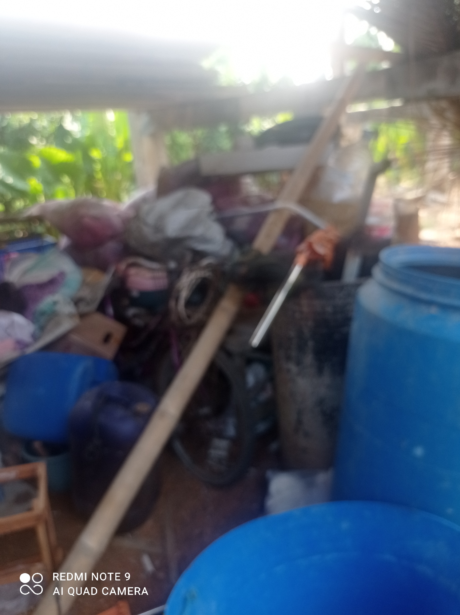 The image depicts an outdoor storage area, likely in a rural setting, with various objects cluttering the space. In the foreground, a large blue plastic container is prominently visible, along with another partially obscured blue container. To the left, a smaller wooden structure is present. The background is filled with a jumble of items including bags of fabric, what appears to be trash or discarded materials, a wooden plank, and a bicycle wheel. The scene is dimly lit, with sunlight streaming from the upper left, creating a bright, hazy effect that obscures details. The environment appears to be somewhat overgrown with vegetation visible in the background, suggesting a natural or semi-natural outdoor space. The presence of large plastic containers and discarded items hints at a potential use for storage or waste management. The image does not contain any people or specific indications of immediate action or interaction. The text "REDMI NOTE 9 AI QUAD CAMERA" is overlaid on the bottom left corner, indicating the device used for capturing the photograph.