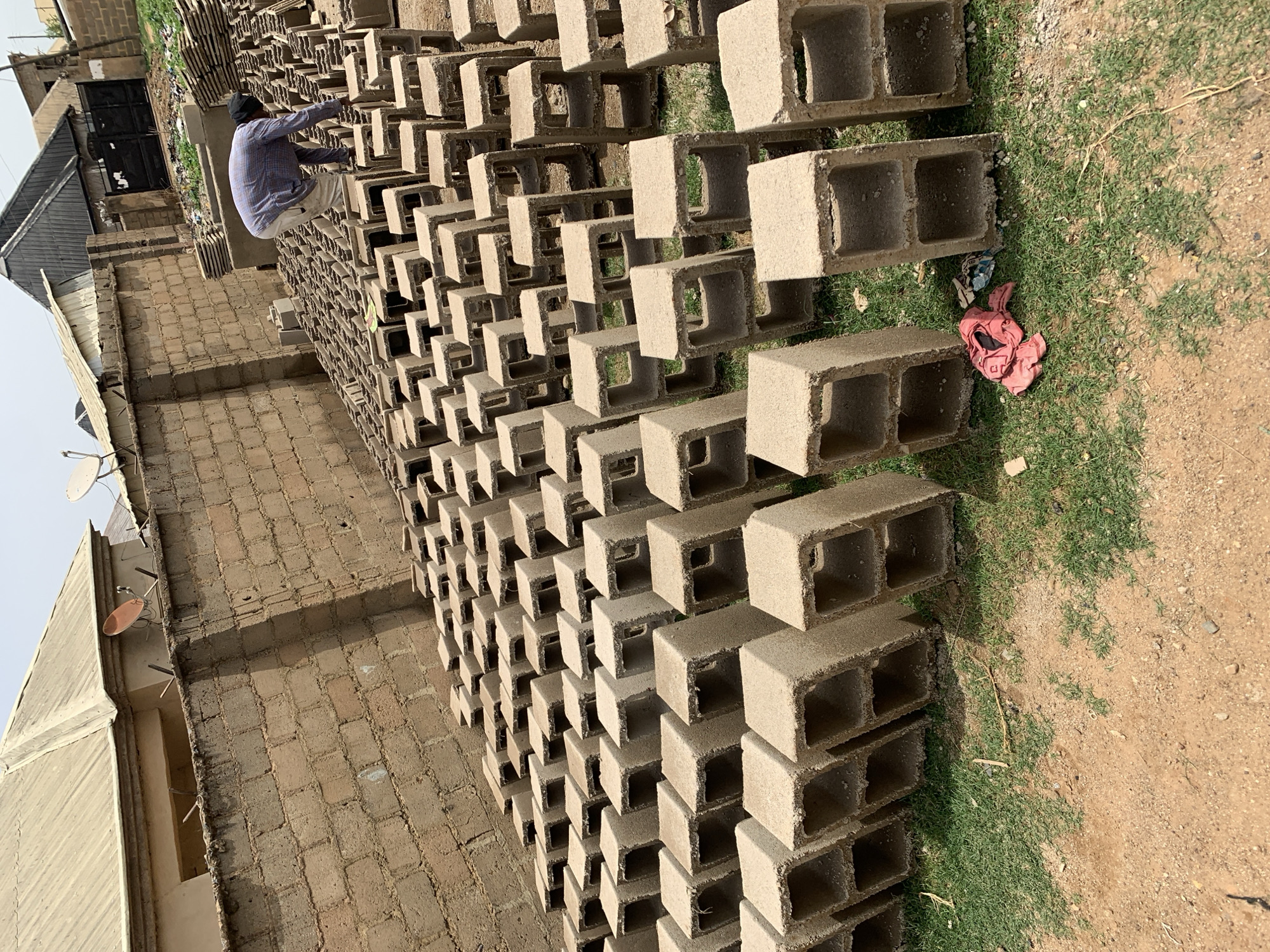 A man wearing a blue shirt and light-colored pants stands atop a large stack of concrete blocks in Rafin Par, Nigeria. The blocks are stacked in neat rows. To the left is a brick building. The ground on the right is partly covered with green grass and bare earth.