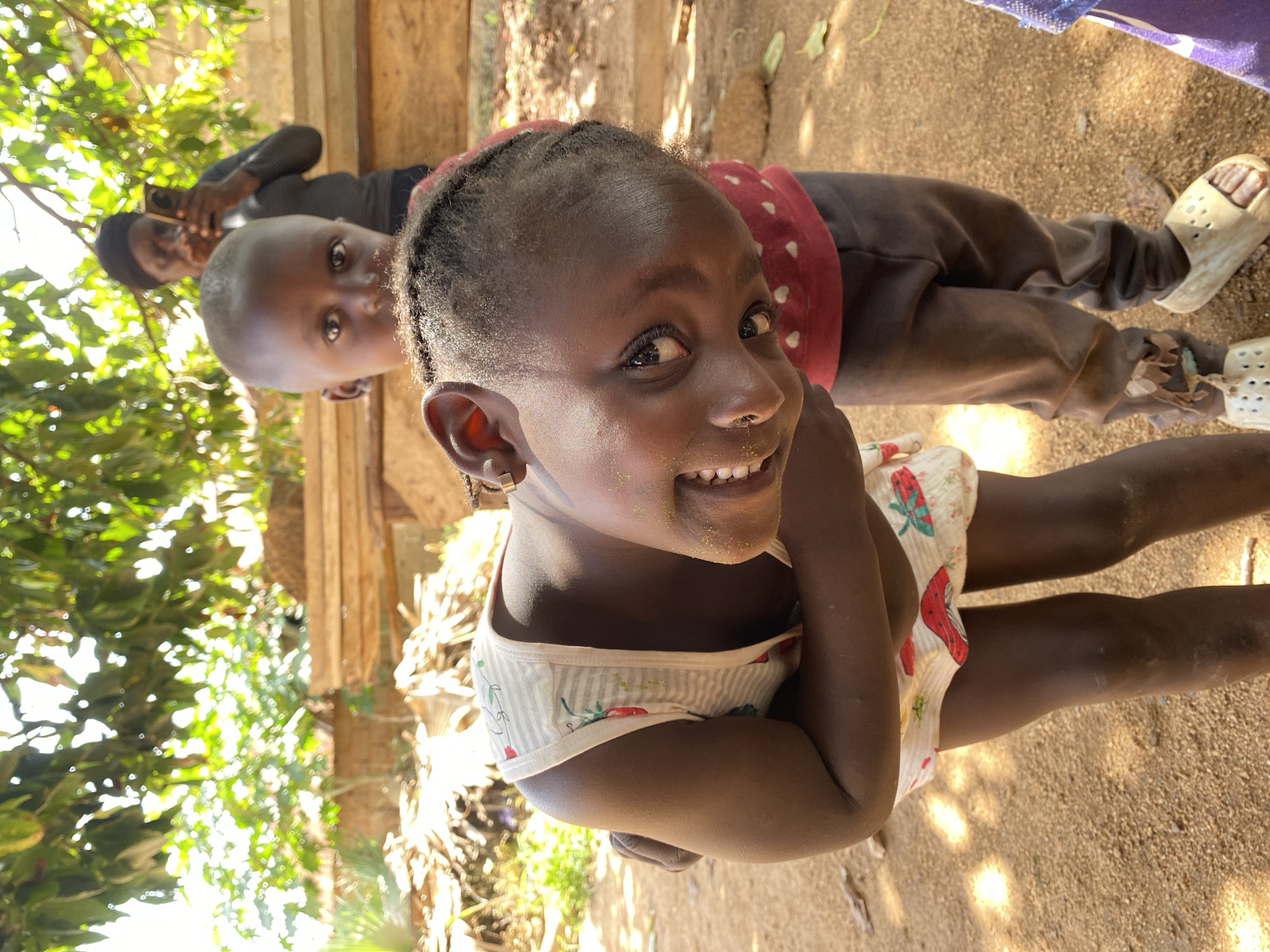 A high-angle, slightly-off-horizontal medium shot captures a young, dark-skinned child smiling directly at the camera. The child wears a light-colored, patterned top with red mushroom designs.
Behind the child, a second, similarly-aged child, dressed in brown pants and a red polka-dot top, is partially visible. In the background, a person is holding a cell phone.
The environment consists of a ground covered in dirt and sand, partially shaded by trees and vegetation on the left side. Some stacked wooden planks are in the background.
Given the provided prompt and the fact that Riyom is a mostly rural area with minimal modern infrastructure, one can infer that the picture was taken in a rural surrounding of Riyom, Nigeria.