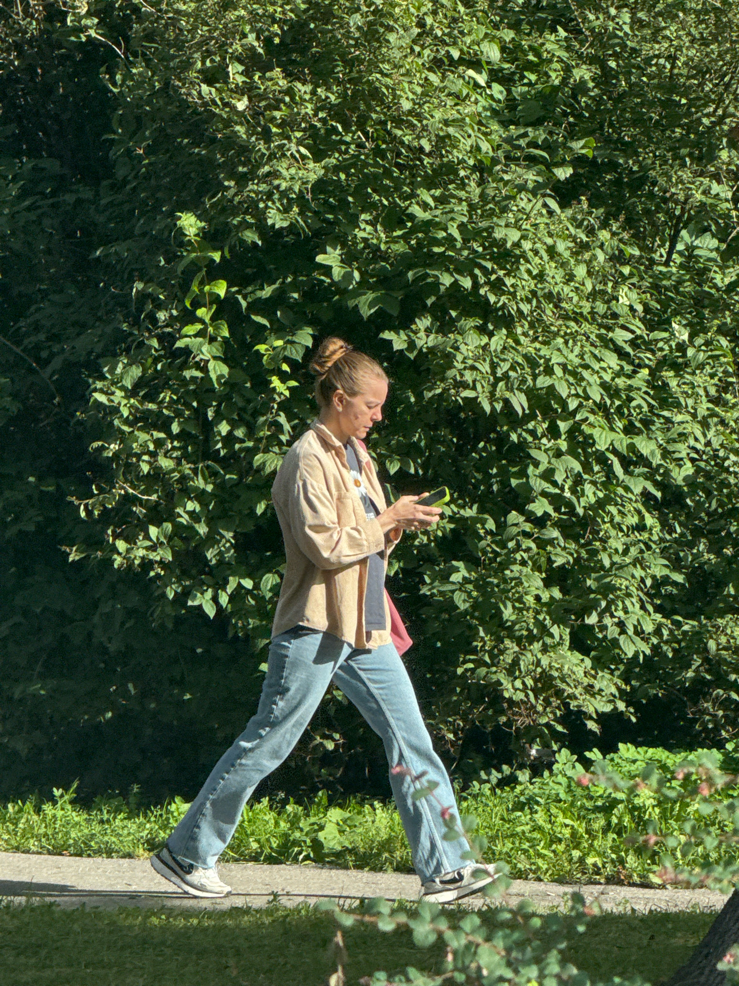 A woman with blonde hair pulled up in a bun walks on a paved path outdoors, looking at her smartphone. She is wearing a light-colored collared shirt, blue jeans, and athletic shoes. A bright pink bag is slung over her shoulder. The background is filled with lush green foliage from trees and bushes, suggesting a park or a treelined street. The lighting indicates it is daytime, with sunlight filtering through the leaves. The scene is likely located in a park within Saint Petersburg, Russia, given the presence of well-maintained greenery and public pathways typical of the city's recreational areas.