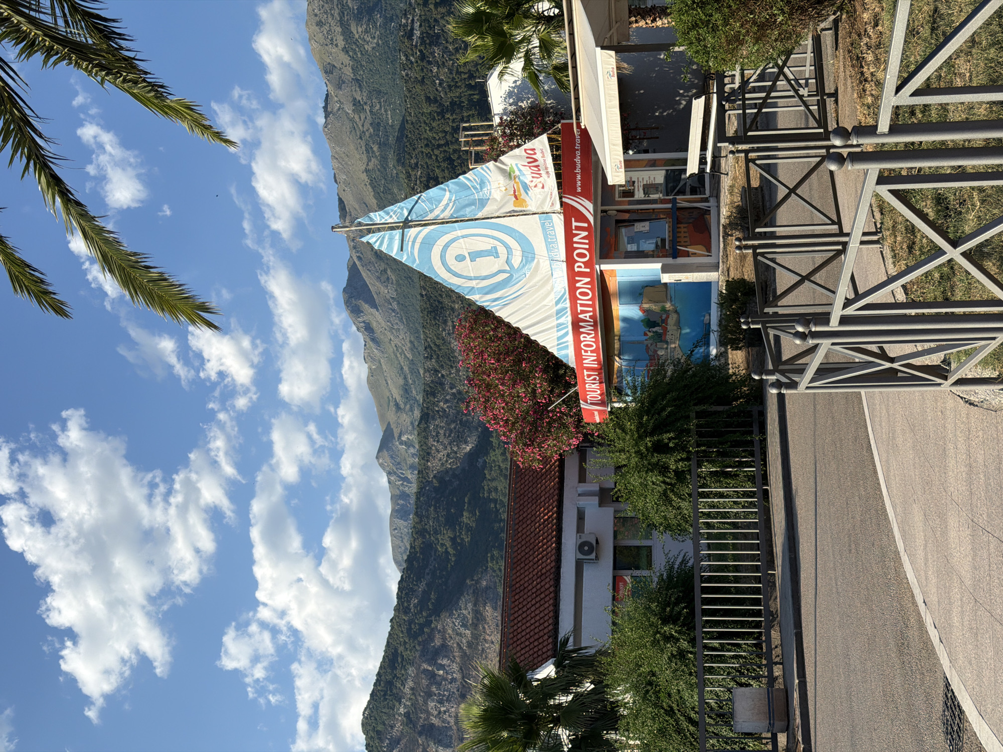 The image depicts a tourist information point designed to resemble a sailboat, located in Budva, Montenegro. The structure features a white sail with a blue outline and a large blue "i" symbol, indicating information. Text on the sail reads "i budva.travel" and "SUDVA." Below the sail, a red banner proclaims "TOURIST INFORMATION POINT." The information point is situated in front of a white building with a red-tiled roof, which has an air conditioning unit visible on its exterior. Green bushes and a palm tree are present around the building and the information point. In the background, a range of green, mountainous terrain rises against a bright blue sky filled with white, fluffy clouds. A metal fence and a paved walkway are also visible in the foreground.
