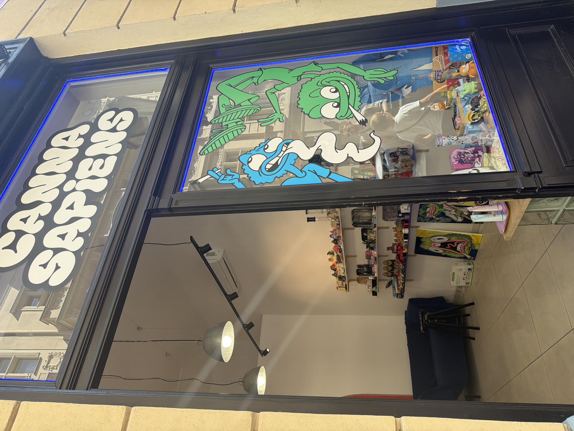 The image shows the storefront of "CANNA SAPIENS" in Prague, Czechia. The storefront features a large glass window with cartoonish illustrations of characters smoking. Inside the store, a blue couch is visible, along with shelves stocked with various products. Two pendant lights hang from the ceiling, and a track light illuminates a section of the interior. The exterior of the building is made of light-colored stone. A reflection of a building across the street is visible in the shop window.