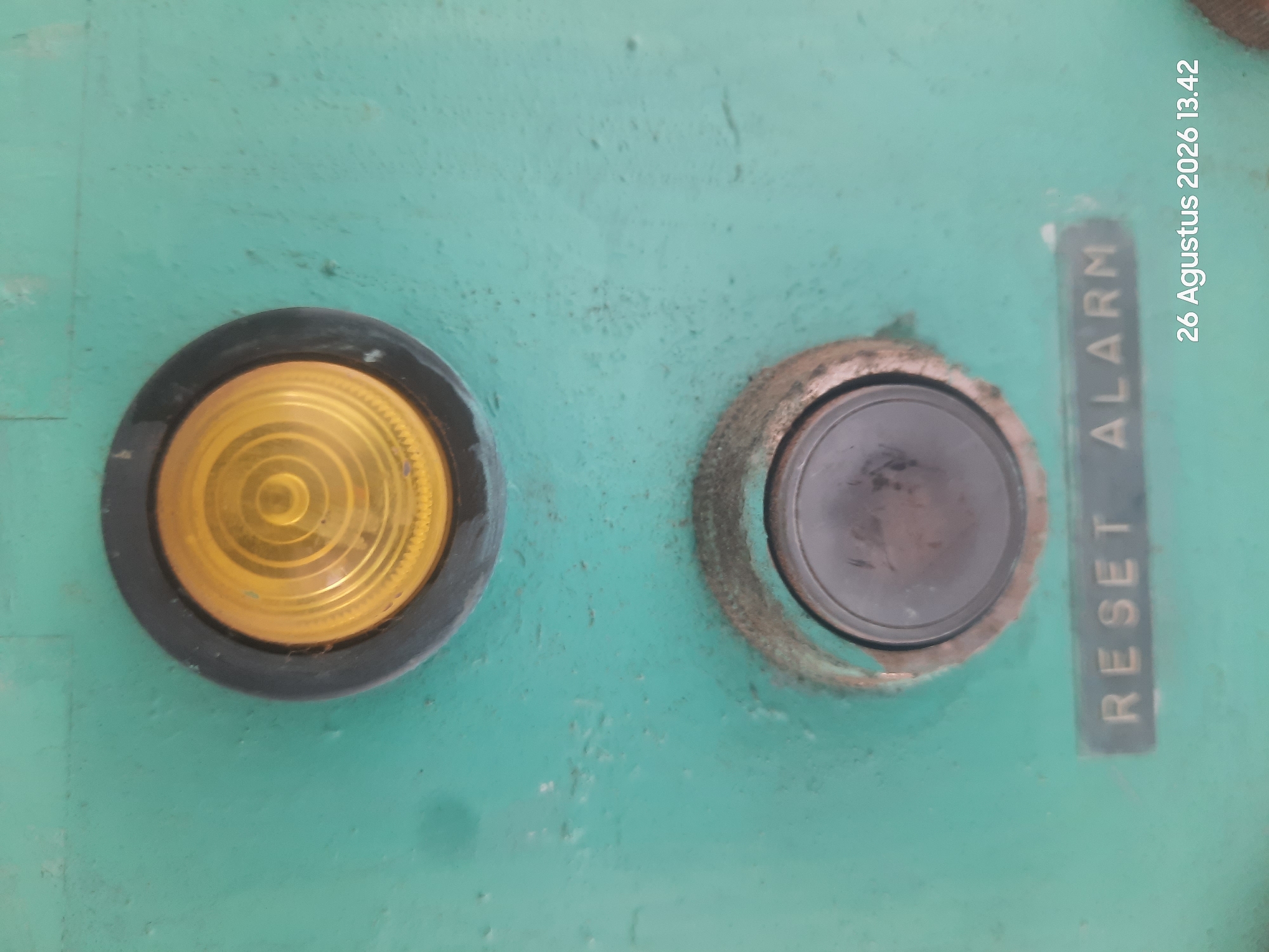 The image shows a close-up view of a teal-colored control panel. At the top of the panel is a round, amber indicator light with a black bezel. Below the indicator light is a round, black button with a black bezel that appears to be a push button. Below the button, there are two labels. The left label reads "RESET" in gold lettering, and the right label reads "ALARM" in gold lettering. The control panel is weathered and shows signs of wear and tear. A timestamp in the lower right corner of the image indicates "26 Agustus 2026 13.42". This image is believed to have been taken in the city of Gending, Indonesia.