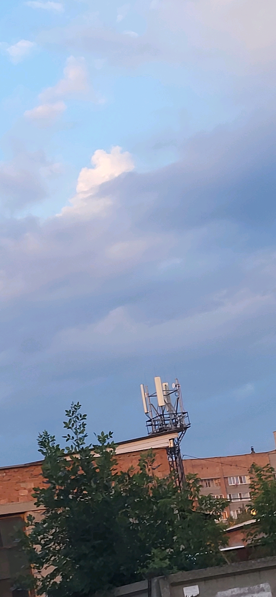 The image displays a communication tower situated on top of a brick building. The tower is equipped with several antennas. In the foreground, a lush green tree partially obscures the view of the building and the tower. The sky above is filled with a mix of light and dark clouds. The scene appears to be an outdoor urban environment, potentially within the city of Oskemen, Kazakhstan, given the context. There are no people visible in the image. The overall impression is one of an overcast sky with industrial infrastructure and natural elements.