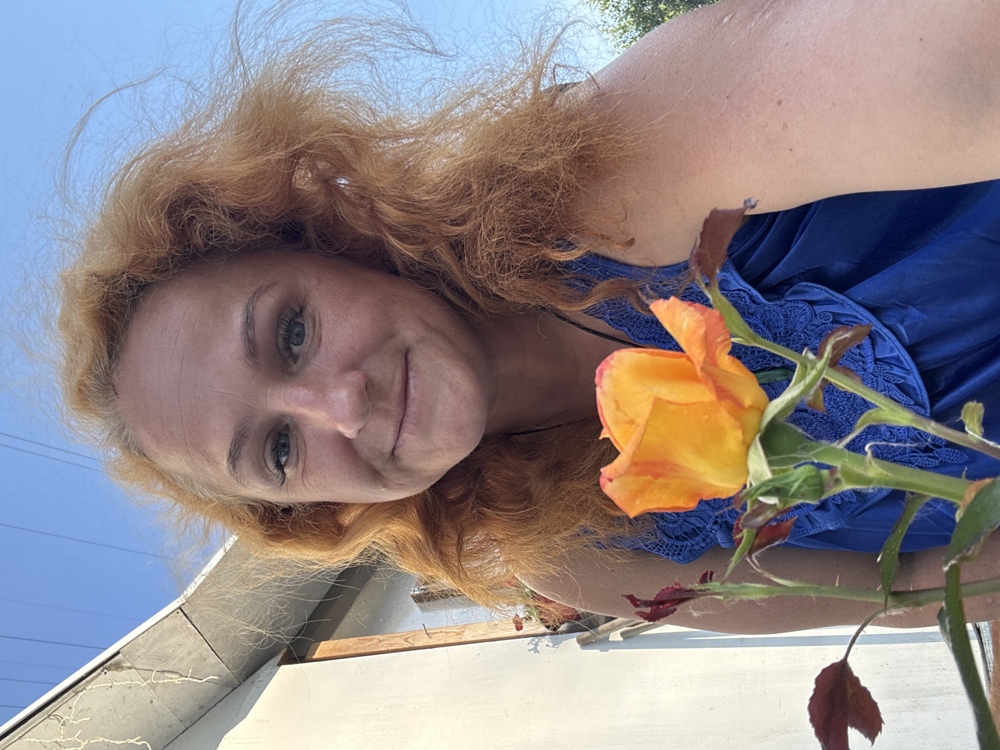 A woman with red hair is holding a yellow and orange rose. She is wearing a blue dress. The woman is standing outside, and there is a blue sky in the background. The scene appears to be in a residential area, with a house visible on the left side of the image. The woman is smiling at the camera.