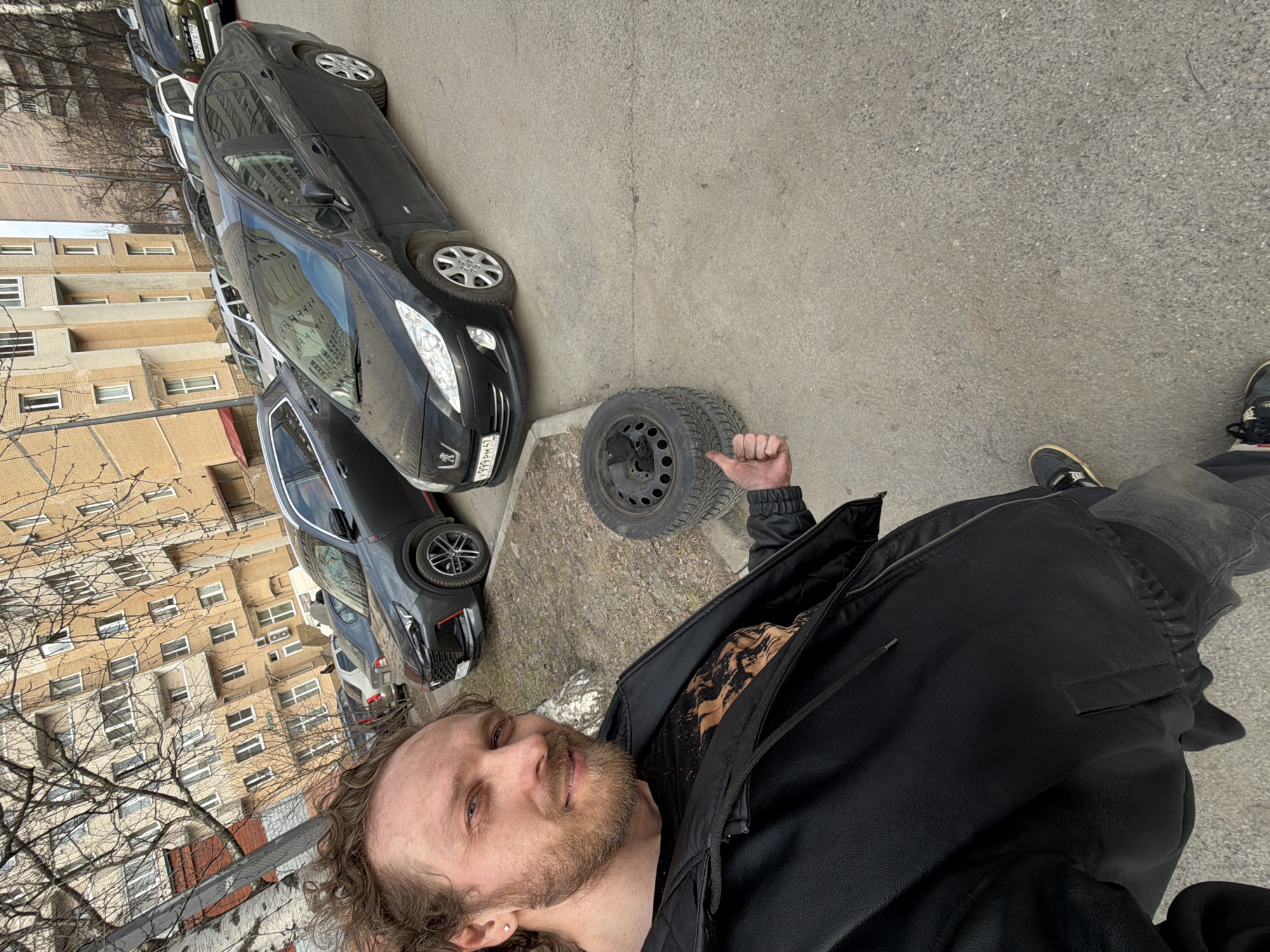 A man in a black jacket and grey jeans stands on a grey pavement next to a car tire, giving a thumbs-up. Behind him are several parked cars, followed by a residential building with many windows. Bare trees are visible in front of the building. The license plate on the car closest to the tire reads "K999PM47". This location is likely in the city of Saint Petersburg, Russia.