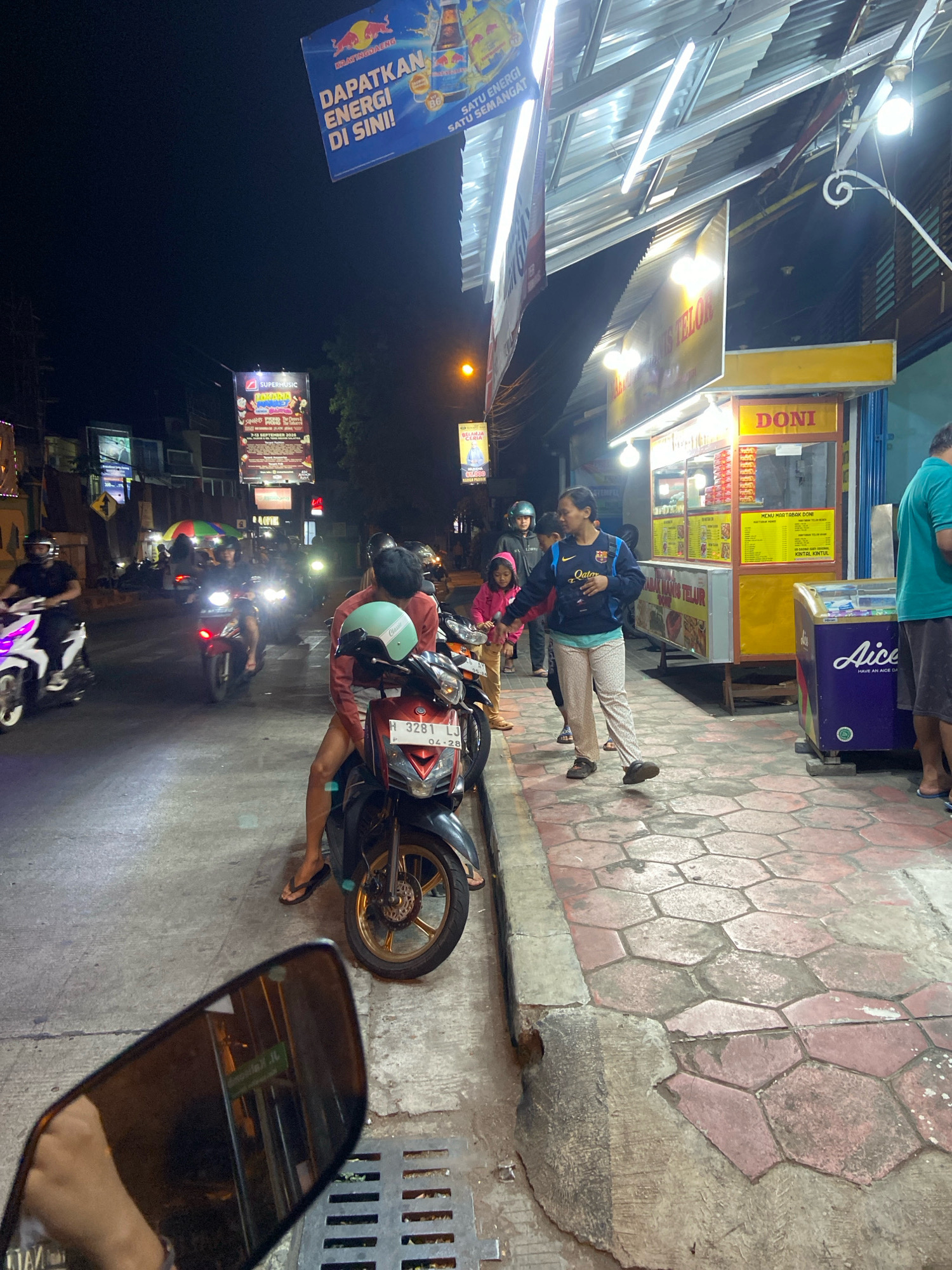The image captures a nighttime scene on a street in Salatiga, Indonesia, likely near a food stall. In the foreground, a motorcycle's rearview mirror reflects a portion of the street and a person's arm. The main focus of the image is a food stall with the name "DONI" displayed prominently in yellow lettering. The stall has a yellow awning and is illuminated by overhead lights. Menu boards are visible, advertising food items. Adjacent to the stall is a purple ice cream freezer with the brand "Aice" visible.
On the sidewalk in front of the stall, several people are present. A woman in a dark jacket and light-colored pants is looking towards the stall, possibly interacting with someone. To her left, a young child in a pink jacket stands near another person. Further down the sidewalk, a man in a green shirt is standing.
The street is bustling with motorcycle traffic, typical of Indonesian urban nights. Several motorcycles are parked or in motion. In the mid-ground, two individuals are seated on a motorcycle, one wearing a red long-sleeved shirt and a light green helmet. Another motorcycle is in motion to the left, ridden by a person wearing a black helmet.
Overhead, a banner for "KRATINGDAENG" is displayed, promoting energy drinks. Signs and streetlights contribute to the overall illumination of the scene. The ground is paved with hexagonal tiles in red and gray. The environment suggests a lively commercial area at night.