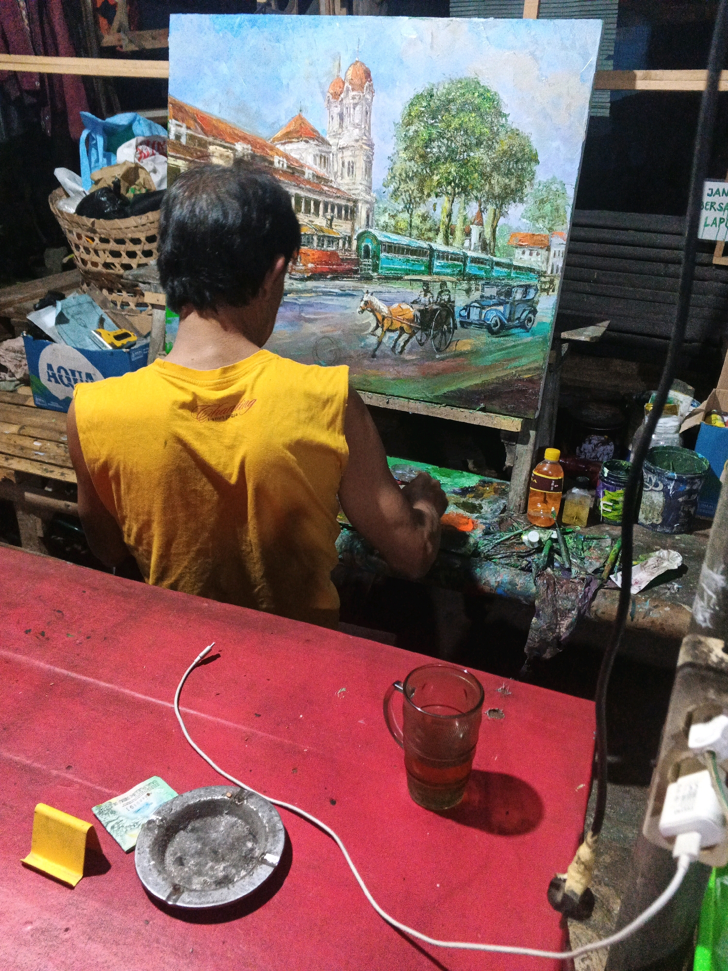 The image depicts a man in a yellow t-shirt painting a landscape scene on a canvas. The painting features a building with orange domes, trees, a horse-drawn carriage, and an old-fashioned car. The man is working at a cluttered table with art supplies, including paint bottles, brushes, and a palette. In the foreground, a red table holds a glass of tea, an ashtray with cigarette butts, a piece of paper money, and a yellow object that appears to be a phone stand. A white cable runs across the table and connects to a power outlet. The background shows a basket filled with various items, a blue box with the "AQUA" logo, and stacked wooden planks. The overall environment suggests an artist's studio or workspace. The signage "JAM BERSAMA LAPUR" is visible on a sign in the background, which could be indicative of a location in Ungaran, Indonesia, possibly relating to a local establishment or event.