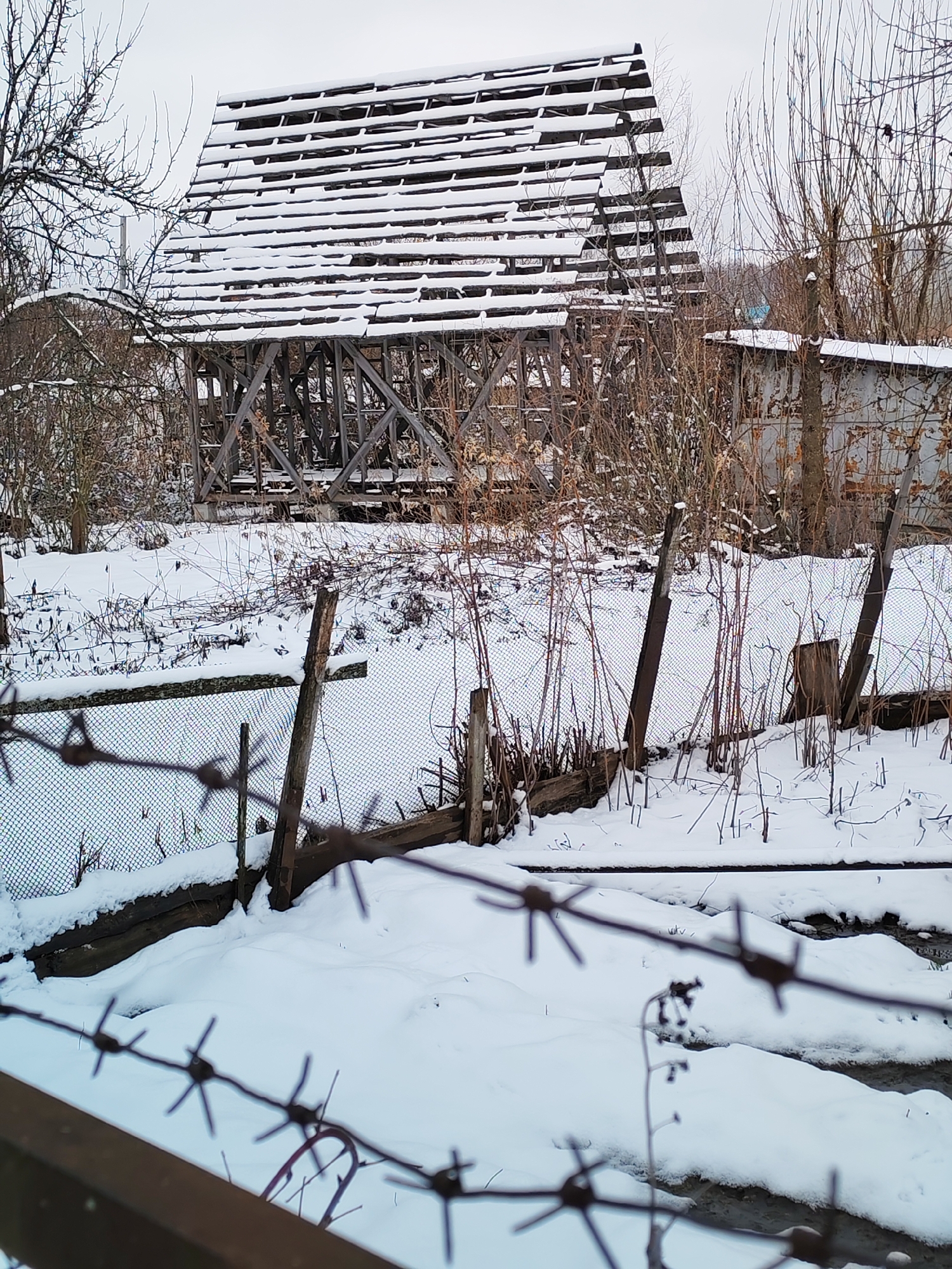 The image depicts a snow-covered scene, likely in a rural or semi-rural area within the city of Chaikovsky, Russia. In the background stands the dilapidated framework of a building, possibly a barn or shed, with a partially constructed roof laden with snow. The structure is made of wooden beams and slats. An adjacent metal building is visible. The foreground features a barbed wire fence, partially obscured by snow, and a frozen or partially frozen stream running through the scene. Leafless trees and shrubs dot the landscape, adding to the wintry atmosphere. There are no people visible in the image.