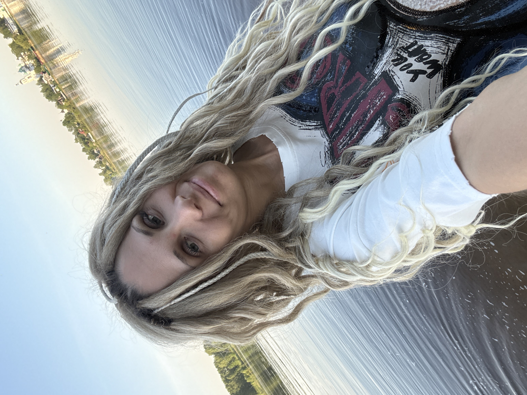 A selfie of a woman with long, wavy blonde hair and braids in her hair, wearing a white t-shirt with a print, is taken near the water in the city of Rusanovo, Russia. The water reflects the trees and buildings of Rusanovo in the background. The sky is visible on the left side of the image.