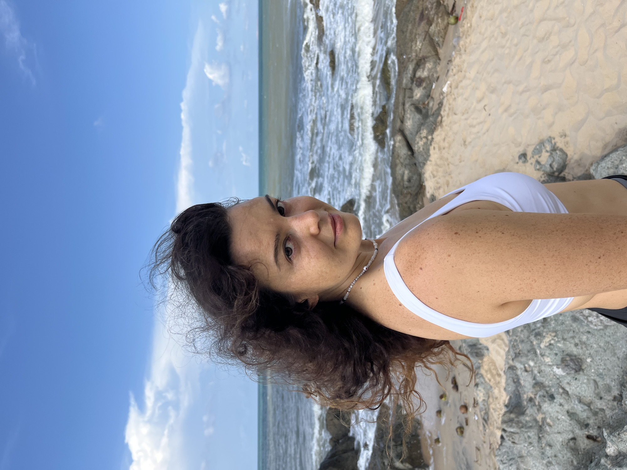 A young woman with dark, curly hair stands on a rocky, sandy beach. She is wearing a white bikini top and a beaded necklace. Her hair is windswept, and she is looking over her shoulder at the camera. The ocean is visible behind her, with waves crashing on the shore. The sky is blue with scattered clouds. The image appears to have been taken in Phu Thuy Ward, Vietnam.