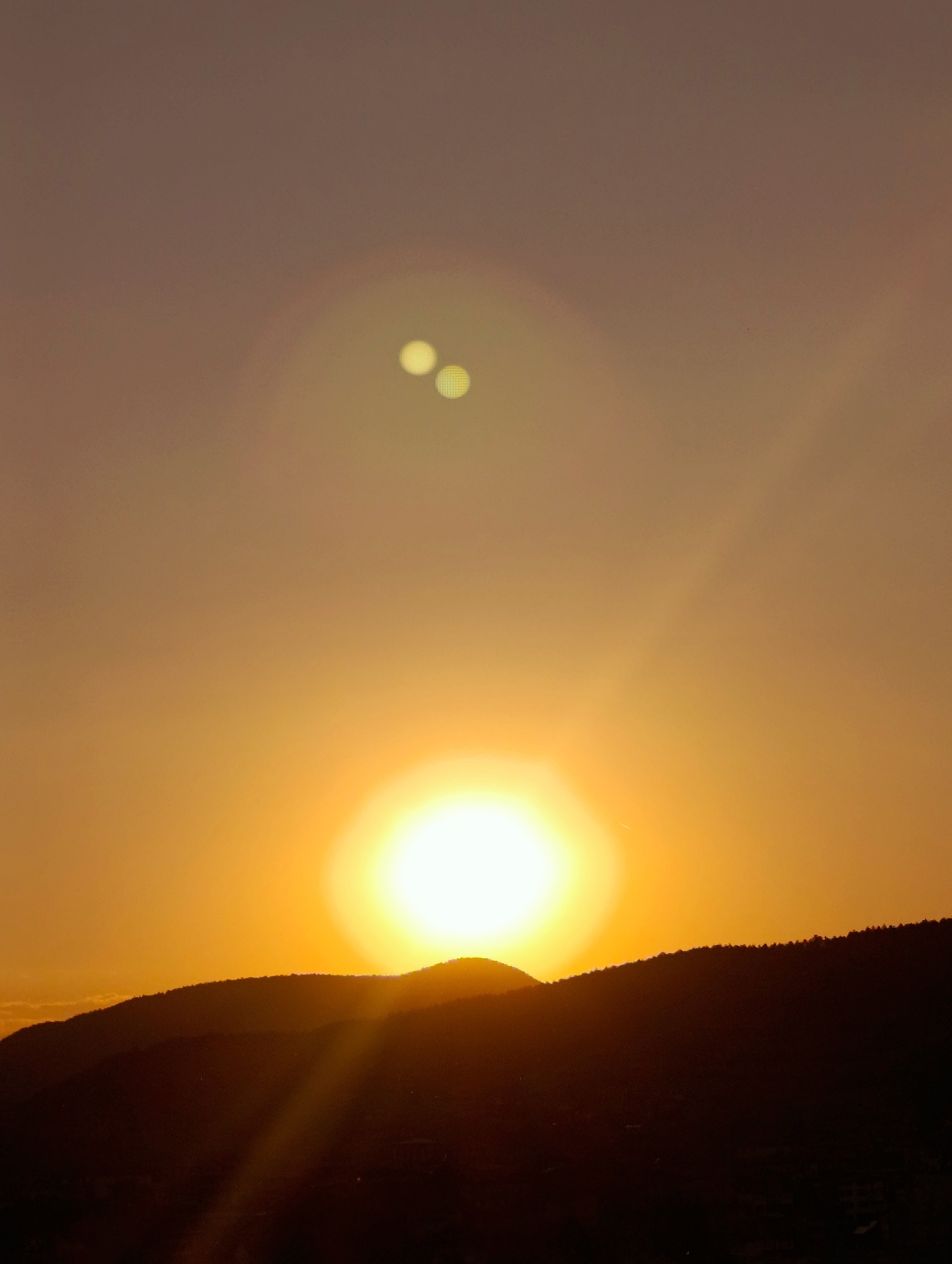 An image captures a sunset over the city of Kardzhali, Bulgaria. The sun, a bright orange orb, is positioned just above the horizon, casting a warm glow across the sky. Silhouetted against the vibrant sky are rolling hills that appear to be covered in trees. The lower portion of the image is dark, obscuring details of the city itself, but hints of lights suggest it is inhabited. The sky is a gradient of warm colors, ranging from deep orange near the sun to a muted purple at the top of the frame. Two circular lens flares, appearing as bright yellow spots, are visible in the upper portion of the sky, indicating the presence of light sources.