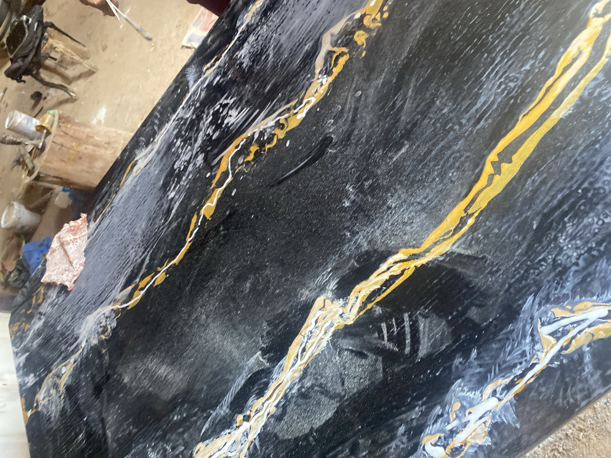 The image depicts a partially finished black countertop with gold and white veining. The countertop is angled, with its top edge visible in the upper right corner and its bottom edge in the lower left. The black background has a textured appearance. On the left, there is a cluttered workbench with cans, tools, and a piece of cloth. The countertop appears to be in a workshop setting. There is no definitive evidence to confirm that this image comes from Jos, Nigeria.