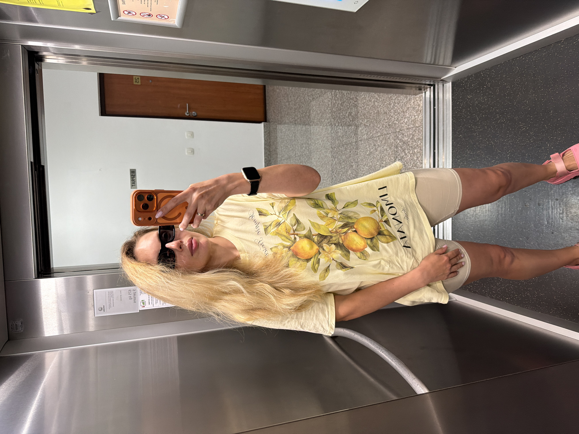 A female individual is captured in a mirror selfie within an elevator located in Budva, Montenegro. She possesses long, blonde, somewhat disheveled hair, and wears large black rectangular sunglasses. Her lips are visible, appearing full. She is dressed in an oversized, light yellow t-shirt featuring a graphic print of oranges or peaches on a branch, with reversed text on the shirt that includes "BUDVA" and "MOMILI". She is also wearing light beige cycling shorts and distinctive pink, chunky flat sandals with a wide strap and velcro closure.
On her right hand, she holds an orange smartphone with a translucent orange case, used to take the selfie; a silver ring is visible on her ring finger, and her fingernails are painted light pink. A black smartwatch with a gold-colored frame is worn on her left wrist.
The elevator's interior walls are constructed of brushed stainless steel, with a dark grey speckled rubber or linoleum floor. A silver horizontal handrail is affixed to the left wall. The open elevator door reflects a hallway beyond, featuring light-colored walls and a similar grey speckled floor. A dark brown wooden door with a silver handle and the reversed number "S2" (indicating '25') is visible in the reflection, along with two white electrical outlets or switches on the adjacent wall.
Various signs are reflected within the elevator: on the left wall, a white sign with reversed text including "АРТИН" and "2 кг" along with "persons 2" (likely capacity information); on the right wall, a yellow warning sign and a framed white sign displaying multiple red crossed-out symbols (e.g., no smoking, no pets) and reversed text, with "ЗЛЕЕТЕ" partially visible. Above the reflection of her phone, reversed text "TAR221" is visible along the top of the elevator door frame.