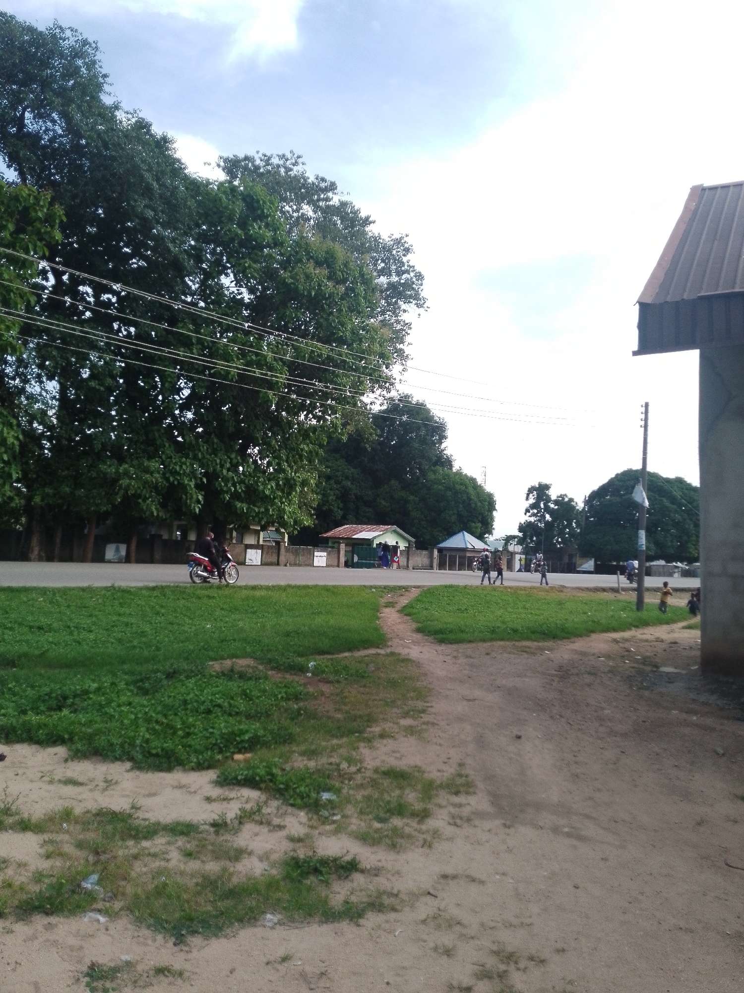 A side view shows a dirt path curving to the right, leading away from a paved road that is bordered by a grassy verge. On the left side of the road, a person on a red motorcycle rides past large, leafy green trees. Beyond the trees, there are several buildings with tiled roofs and walls. On the right side of the dirt path, a portion of a building with a corrugated metal roof is visible. Several people, including some children, are walking and standing near the road and buildings on the right. The sky is bright with some clouds.
