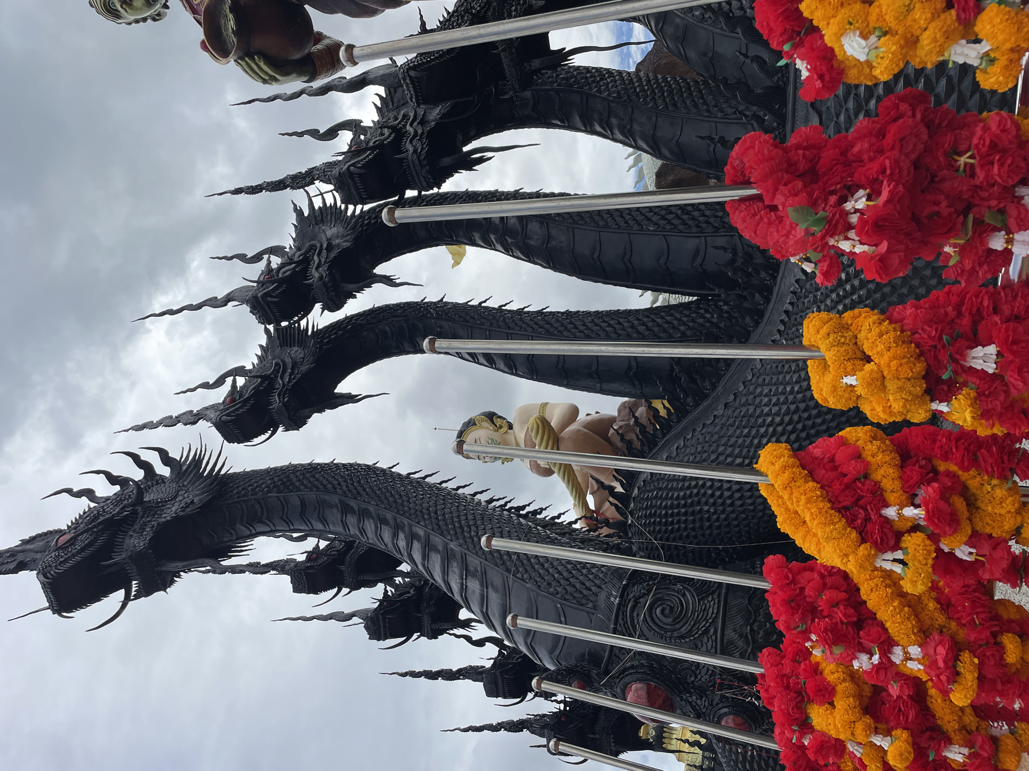 The image shows a large, black sculpture of a multi-headed Naga, a mythical serpent, in Bo Win Subdistrict, Thailand. The Naga has seven heads visible, each with sharp teeth and spiky crests. A statue of a serene figure, possibly a deity or a mythical being, sits on the Naga's body. The sculpture is adorned with garlands of red and yellow flowers in the foreground. The background consists of a cloudy sky. Several metal poles are visible in front of the sculpture, partially obscuring it. The overall scene is imposing and likely part of a religious or cultural site.
