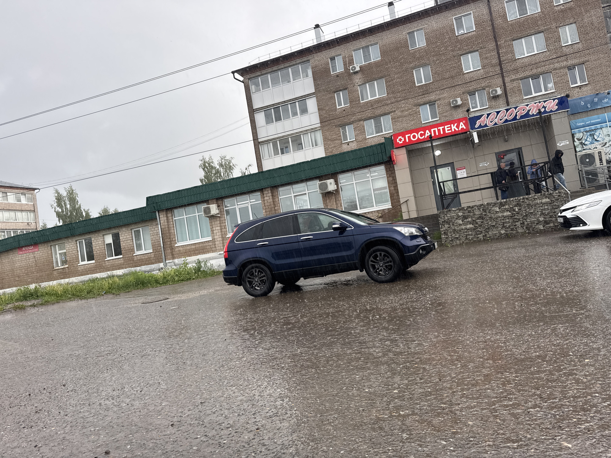 The image depicts an urban street scene in Mozhga, Russia, on a rainy, overcast day. The paved ground is visibly wet, covered in puddles with numerous ripples indicating ongoing rainfall.
A dark blue Honda CR-V SUV is positioned in the midground, angled towards the left, with its black wheels prominent. The front of a white car is partially visible on the far right.
Two brick buildings form the backdrop. To the left, a lower, elongated building with multiple windows and a dark green roof features a small "АРЕНДА" (Rent) sign on its side. To the right, a taller, multi-story brick building with numerous windows and external air conditioning units dominates the upper-midground. Its ground floor houses commercial establishments.
A storefront entrance is visible near the center-right, sheltered by an overhang. Above the entrance are two illuminated signs: a red one displaying "ГОСАПТЕКА" (Gosapteka - State Pharmacy) with "8 - 20" (likely operating hours) beneath it, and a blue sign that reads "АССОРТИ" (Assorti - Assortment) adorned with decorative lights. Several people are gathered near this entrance, possibly sheltering from the rain. One person wears a blue hooded jacket, another a dark one. To the far right, a poster for "Товары для пикника" (Goods for picnic) is visible, along with a "ZANUSSI" label on an air conditioning unit.
The sky is a uniform, heavy grey, indicative of the rainy weather. Overhead, several power lines traverse the frame. The scene conveys a typical, mundane rainy day in an Eastern European urban setting.