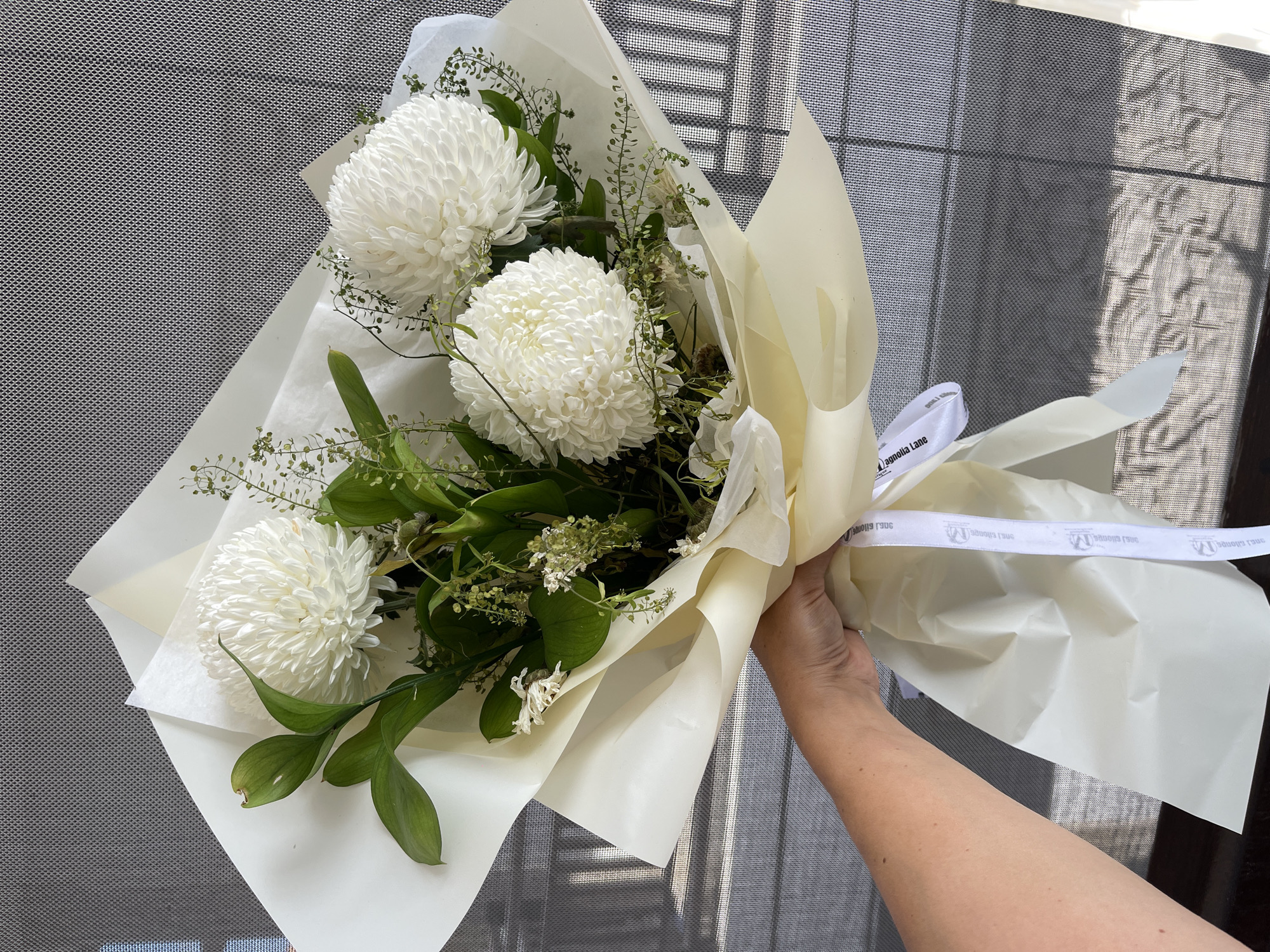 A bouquet of white chrysanthemums, green leaves, and delicate greenery is held by a person's hand, wrapped in pale yellow and white paper. The bouquet is arranged with the flowers facing upwards, and a white ribbon with black lettering is tied around the lower part of the wrapping paper. The background consists of a grey, patterned surface that appears to be a mesh or screen. Shadows are cast on the background, suggesting the presence of light. The image appears to have been taken outdoors, possibly on a balcony or near a window, as evidenced by the light and shadows. The city of Kuta, Indonesia is not depicted in the image.