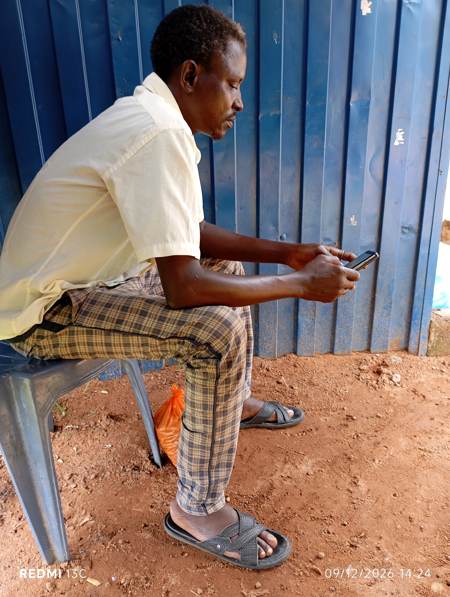 A man is sitting on a blue plastic chair outdoors, facing left. He is wearing a light-colored, short-sleeved collared shirt and plaid pants. The man is holding a black mobile phone in his hands and appears to be looking at it. The background consists of a blue corrugated metal wall. The ground is covered with reddish-brown dirt. To the right of the chair, an orange plastic bag is visible on the ground. A timestamp in the lower right corner indicates "09/12/2026 14:24". The location is identified as Shendam, Nigeria.