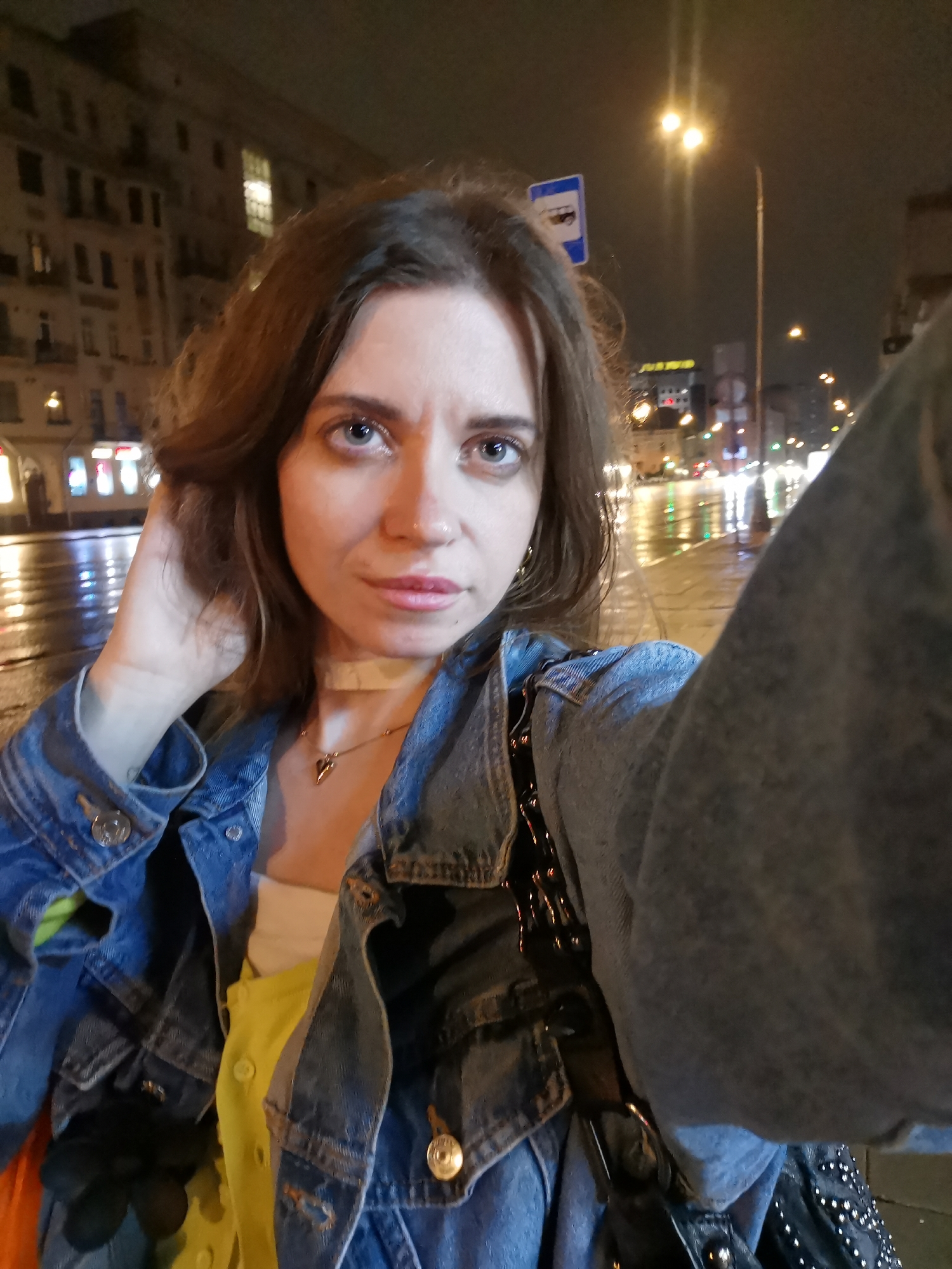 A woman is taking a selfie at night on a wet city street. She is wearing a denim jacket over a yellow shirt and a necklace. Her hair is brown and her eyes are blue. The background shows city buildings, street lights, and a bus stop sign. The city is wet with reflections from the lights.