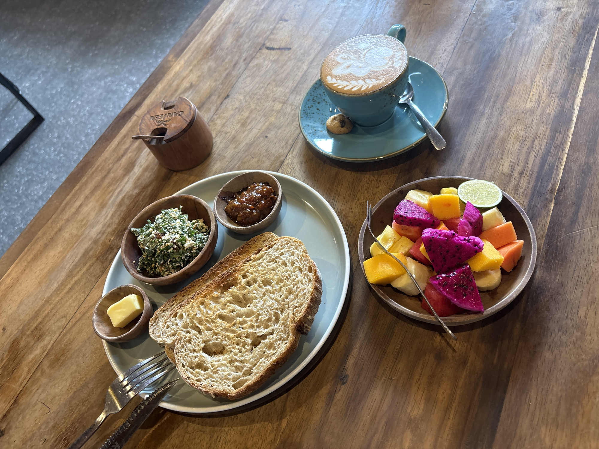 The image depicts a breakfast scene at a location in North Kuta, Indonesia. The scene includes a wooden table with a variety of food items:
* A plate with a slice of toasted bread, a wooden bowl containing a green salad, another bowl containing a red-colored relish, and a small bowl with a pat of butter.
* A wooden pot with an inscription (possibly the restaurant name or brand) and a small spoon.
* A turquoise cup and saucer with a latte art and a small cookie next to it.
* A wooden bowl with a mixed fruit salad containing pieces of banana, dragon fruit, mango, papaya, and a slice of lime.
The utensils include a knife and fork on the plate with the toast and a spoon on the saucer next to the coffee cup, and a fork in the fruit salad bowl. The environment appears to be a restaurant or cafe setting, with a portion of a chair visible in the top left of the image.