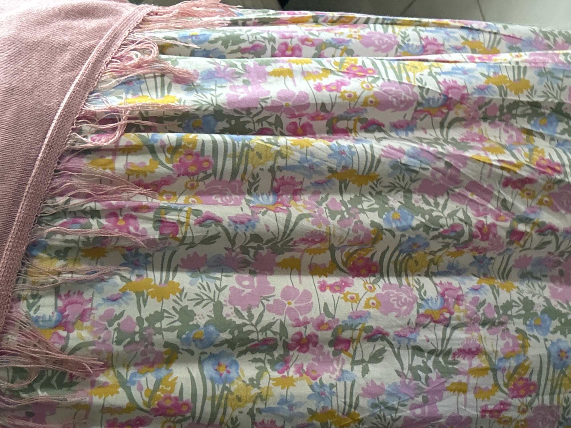 The image shows a close-up of a floral patterned fabric with a pink knitted top with fringe hanging over it. The fabric is a light color with an all-over print of flowers and leaves in shades of pink, yellow, and blue, with green stems and leaves. The knitted top is a dusty rose color and has a band of delicate pink fringe along the bottom edge, which drapes over the floral fabric. The fabric is gathered, creating vertical folds and texture. The lighting appears to be natural, coming from the right side of the image, casting subtle shadows and highlighting the textures of both the fabric and the knit. The background is out of focus and appears to be a plain wall or surface. There are no people or identifiable landmarks present to indicate a specific location within Reuleut Timu, Indonesia.
