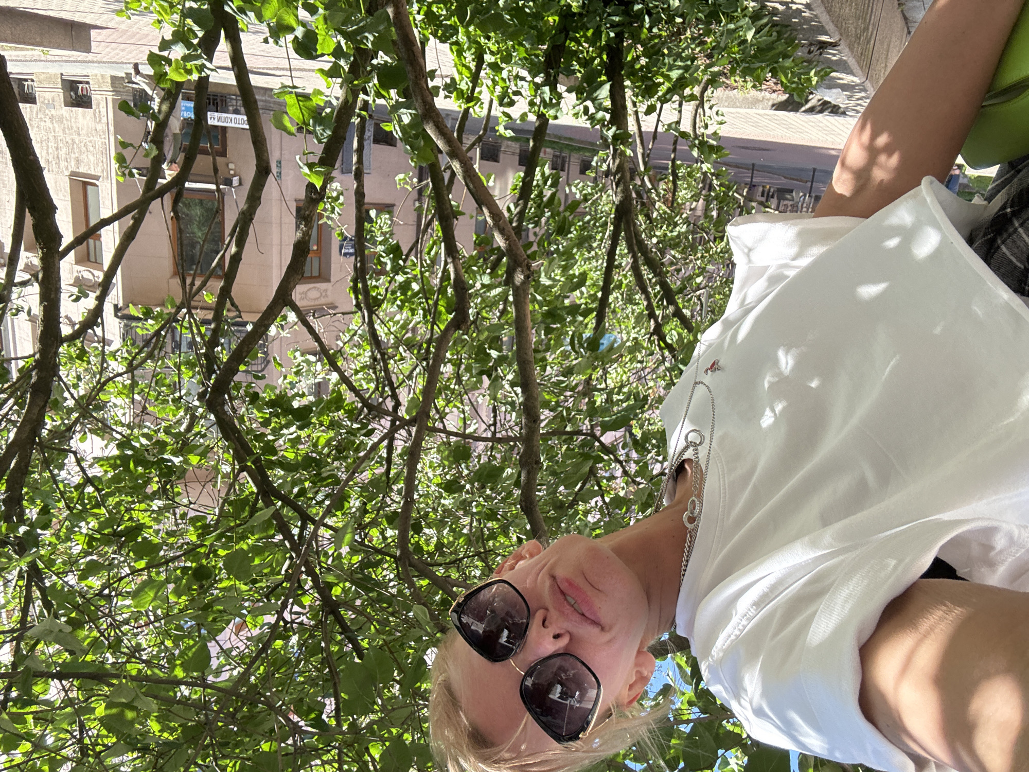The media file is a portrait of a young woman taken from a low angle, looking up at the camera. She is wearing sunglasses, a white t-shirt, and a silver chain necklace. She is outdoors, surrounded by the lush green foliage of trees, with sunlight dappling through the leaves. In the background, a building with visible windows and balconies can be seen. The image is taken in Saint Petersburg, Russia. The time of day appears to be daytime, with bright sunlight and clear skies. The woman's facial expression is relaxed and playful, suggesting a casual and joyful moment. No visible text is present in the image.