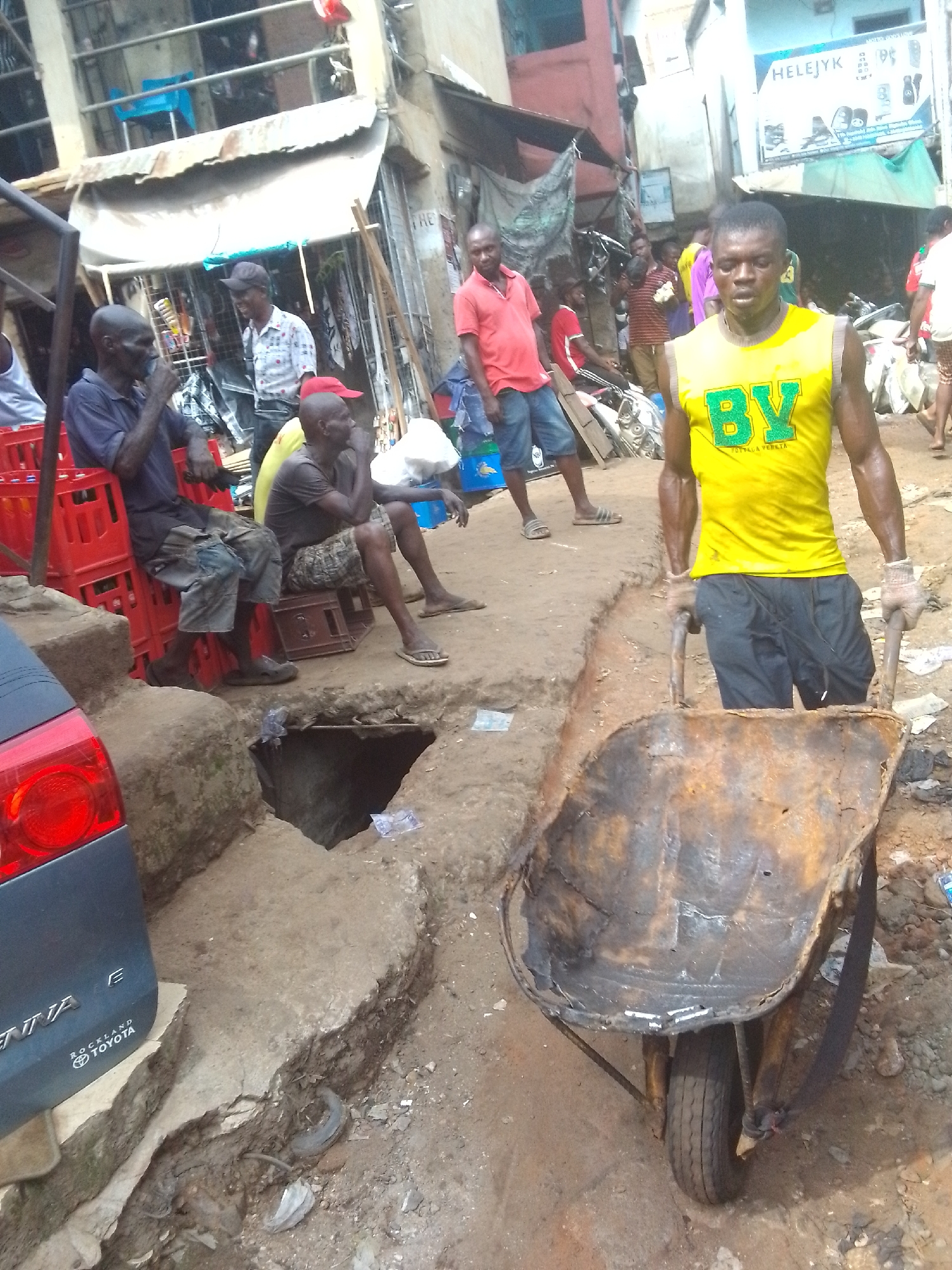 This image captures a bustling street scene in Onitsha, Nigeria, featuring several individuals and makeshift urban infrastructure. In the foreground, a dark-skinned man with a muscular build, wearing a bright yellow sleeveless shirt and dark shorts, pushes a heavily rusted and worn metal wheelbarrow. His yellow shirt prominently displays "BV" in green, with smaller text underneath that appears to read "BOTTEGA VENETA." He wears light-colored work gloves and appears focused on maneuvering the wheelbarrow.
The ground is uneven, composed of packed dirt and broken concrete, with a significant open drain or hole in the immediate path of the wheelbarrow, revealing the challenging conditions of the environment. To the left, the rear section of a dark grey vehicle, identified as a "TOYOTA SIENNA E" with "ROCKLAND" branding, is parked on a raised concrete slab.
Further back on the left, several men are gathered. One man in a dark blue short-sleeved shirt and light denim shorts sits on stacked red plastic crates, appearing to rest or engage in conversation. Another man in a similar position is partially visible next to him. In the mid-ground, other people are seen standing or moving, including a man in a red polo shirt on the left side.
The background consists of various buildings, some with corrugated metal roofs and improvised structures, suggesting a market or commercial area. One building features a blue sign with "HELEJYK" prominently displayed, along with images of electronic devices. The scene is brightly lit, indicating daytime and likely sunny weather. The overall impression is one of a vibrant, working-class urban environment characterized by manual labor and informal gatherings.
