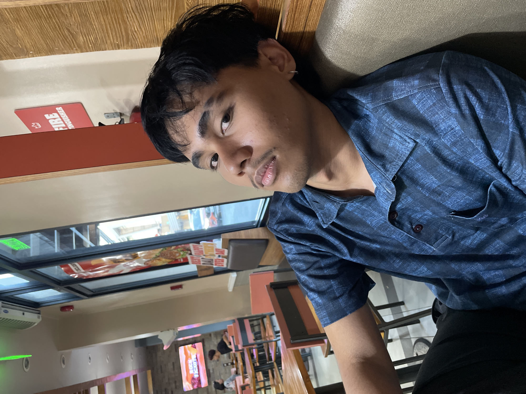 A young Filipino male is seated inside a Burger King restaurant in Davao City, Philippines. He is wearing a dark blue plaid short-sleeved shirt and black pants. The background shows the interior of the restaurant, including tables, chairs, and a window with a view of the street. A red "FIRE EXTINGUISHER" sign is visible on the wall behind him. The lighting is dim, with a greenish light emanating from a fixture above and to the left.