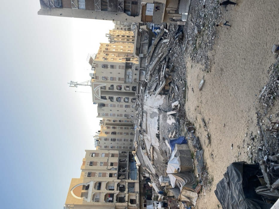 The image portrays a severely damaged urban landscape. Buildings show signs of extensive destruction, with multiple floors collapsed and facades scarred. Debris and rubble are scattered across the ground. In the foreground, makeshift shelters or tents are visible, suggesting displaced people. A sandy area lies adjacent to the damaged buildings, possibly indicating a coastal location. Two individuals are walking in the distance. The overall impression is one of war or disaster.