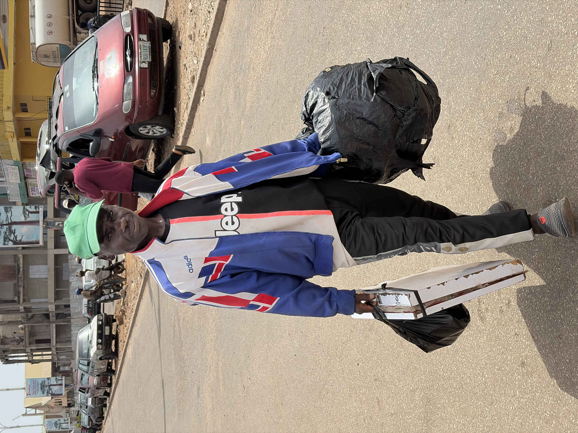 A Black man wearing a green cap, a white and blue Adidas jacket over a black t-shirt with "Jeep" written on it, and black track pants walks on a paved road in Jos, Nigeria. He carries a large black trash bag in his right hand and a white object wrapped in plastic with a black trash bag in his left hand. He is walking away from the camera. In the background, a line of parked cars and buildings are visible. A red van with its doors open is parked on the right side of the road. A water tanker truck is visible in the distance on the right. The sky is bright and overcast.