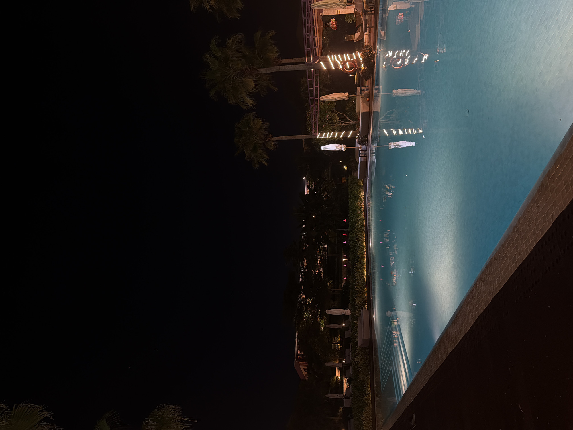 This image presents a nighttime scene at a resort or hotel property located in Dong Ninh Hoa Ward, Vietnam. The central focus is a large swimming pool, illuminated from within, occupying the lower half and center of the frame. The pool water exhibits a light blue hue, reflecting the surrounding artificial lights and structures. A dark tiled border runs along the pool's edge in the foreground, adjacent to a darker, possibly paved or wooden, deck area.
Along the perimeter of the pool, a landscaped area features low-lying bushes with subtle ground lighting. White, folded beach umbrellas are regularly spaced, accompanied by multiple lounge chairs. On the right side of the pool, two tall palm trees are prominent, their trunks adorned with spiraling string lights. Behind these trees, a pergola-like structure is also decorated with string lights, and a red life preserver ring hangs from it.
In the background, multiple multi-story buildings, consistent with a resort complex, are visible. These structures are dimly illuminated by warm-toned interior and exterior lights. Various other trees and dense vegetation are situated between the pool area and the buildings, largely appearing as silhouettes against the faint ambient light. The sky is dark and appears featureless black, with two minute, faint light points discernible in the upper left quadrant. No individuals are present within the depicted scene.