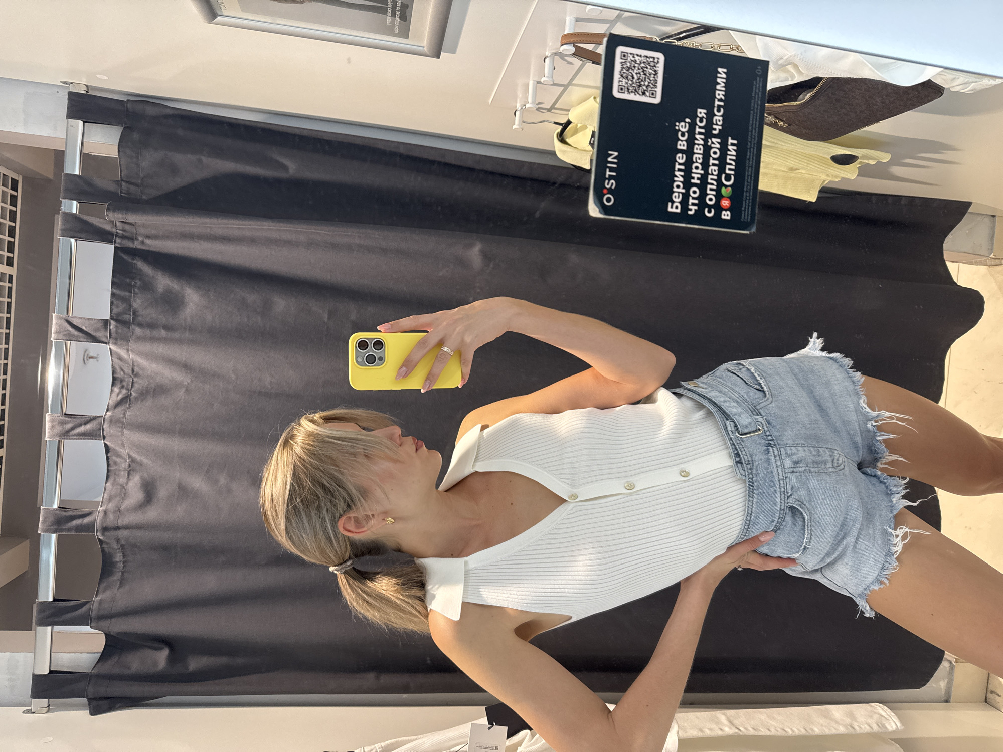 A woman is standing in a dressing room, holding a yellow smartphone in her right hand, taking a selfie in the mirror. She is wearing a white button-down shirt with short sleeves and light blue denim shorts. The dressing room has a black curtain behind her. A promotional sign for the clothing brand "O'STIN" with a QR code is visible near the top right corner of the frame. Various clothes and hangers are visible above the curtain.