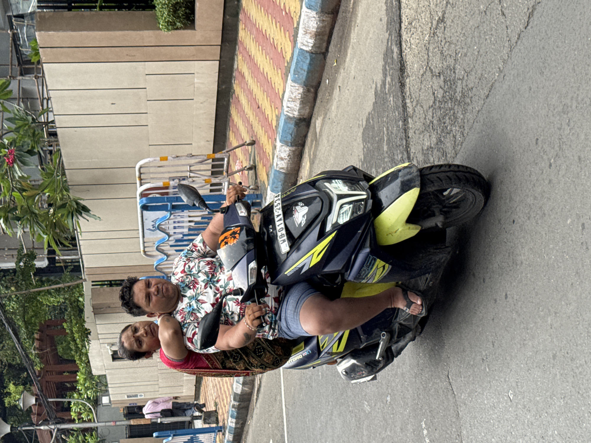 The image, rotated 90 degrees clockwise from its natural orientation, shows two individuals, a man and a woman, on a black and neon yellow TVS NTorq 125 scooter on an urban street. The man, driving the scooter, wears a white shirt with a colorful floral pattern and blue shorts, with his hands on the handlebars. The woman is seated behind him, wearing a red top or saree. Both subjects are looking towards the camera with neutral expressions.
The scooter's front features a license plate displaying "WB 01 AY 2534," indicating its registration in West Bengal, India. The model name "Ntorq 125" is visible on the scooter's front panel.
The scene is set on a clear day, likely daytime, with diffused bright light. The background features buildings, lush green foliage, and various street infrastructure, including blue and white striped barricades. To the side of the road, there is a sidewalk paved with distinctive yellow and red patterned tiles, bordered by a blue and white painted curb, which is characteristic of Kolkata, India. The road itself is asphalt. The overall activity depicted is a common street scene of people commuting or traveling on a scooter in an Indian city.