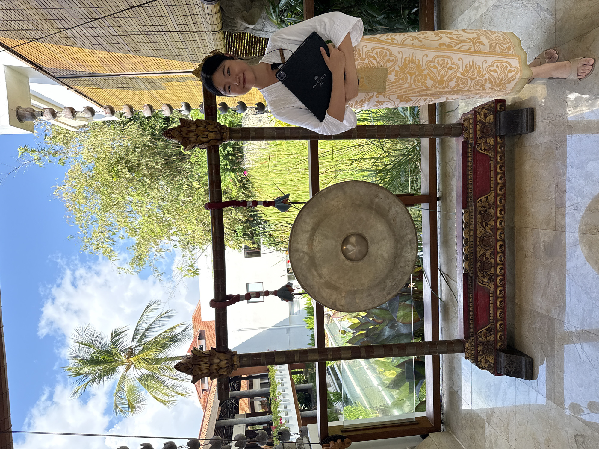 A young East Asian woman is captured in a bright, open-air setting, likely a resort in Kuta Selatan, Indonesia. She stands and smiles gently towards the camera, dressed in a white long-sleeved top and a light-colored sarong or skirt adorned with a prominent golden patterned design. She wears light-colored sandals on her feet. In her arms, she holds a black tablet or laptop case horizontally, on which the white text "PARADISUS BALI" is clearly visible.
To her right, a large, circular metal gong hangs from an intricately carved wooden stand. The stand features traditional red and gold decorative elements. A mallet with a red and white patterned ribbon hangs from the stand. The background is vibrant with lush tropical greenery, including bamboo-like plants and a prominent palm tree reaching into a bright blue sky scattered with white clouds. Modern buildings with white facades and some reddish-brown tiled roofs are visible amidst the foliage.
Above, a bamboo-slat ceiling or awning can be seen, from which a string of decorative, bell-shaped elements is suspended. The ground is a highly reflective, polished tiled floor, likely marble, which mirrors the daylight and parts of the surroundings. The scene depicts a sunny day, suggesting a pleasant, relaxed atmosphere at a tropical destination.