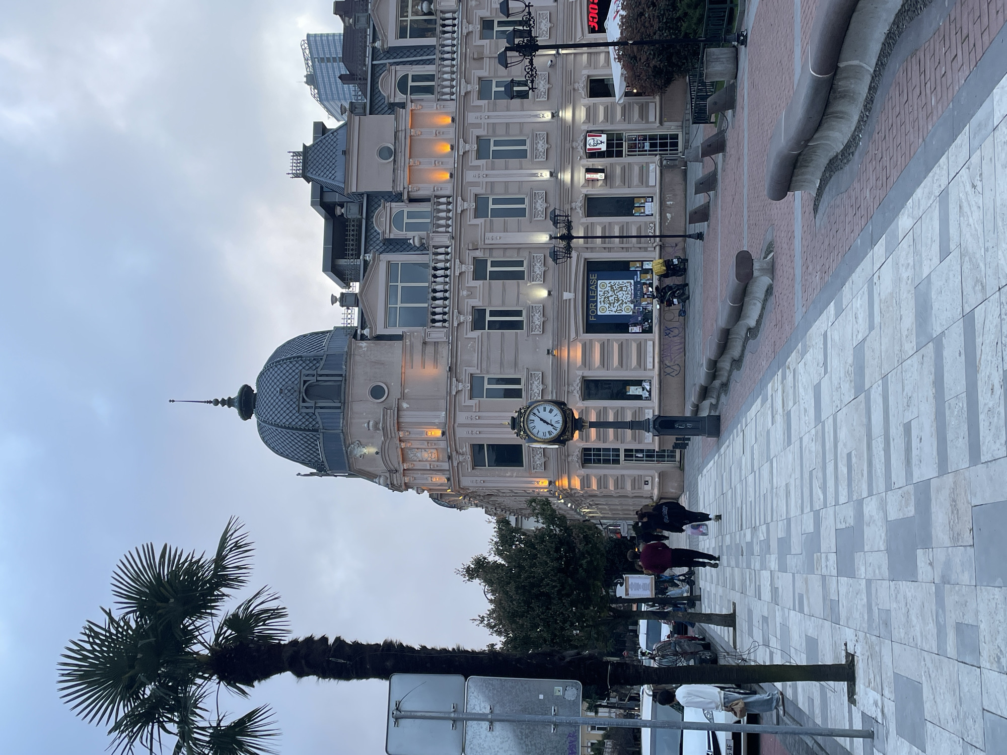 The image depicts a street scene in Batumi, Georgia, featuring the astronomical clock tower building. The building has a dome-shaped roof with a spire. There are multiple levels to the building with windows and architectural details, and a clock is positioned on a pole in front of the building. Pedestrians are walking on a patterned sidewalk. A palm tree is visible on the left, and street signs are also present. The sky is overcast. A KFC restaurant is visible on the building. A QR code for real estate is on display.