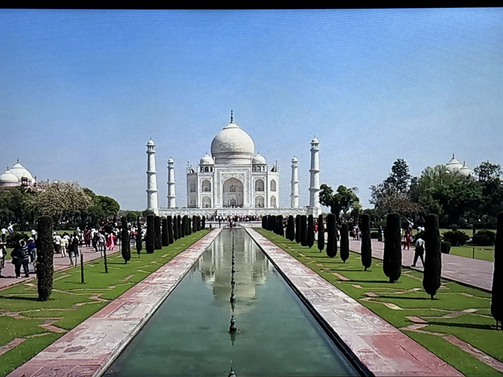 The image shows the Taj Mahal in Agra, India. The iconic white marble mausoleum stands prominently in the center, with its dome and minarets visible against a clear blue sky. A long, rectangular pool stretches from the foreground towards the structure, reflecting the Taj Mahal in its waters. The pool is flanked by well-manicured lawns and rows of dark green, cylindrical trees. Groups of people can be seen walking along pathways on either side of the pool. Another building complex sits to the right of the Taj Mahal, across a green lawn and some trees.