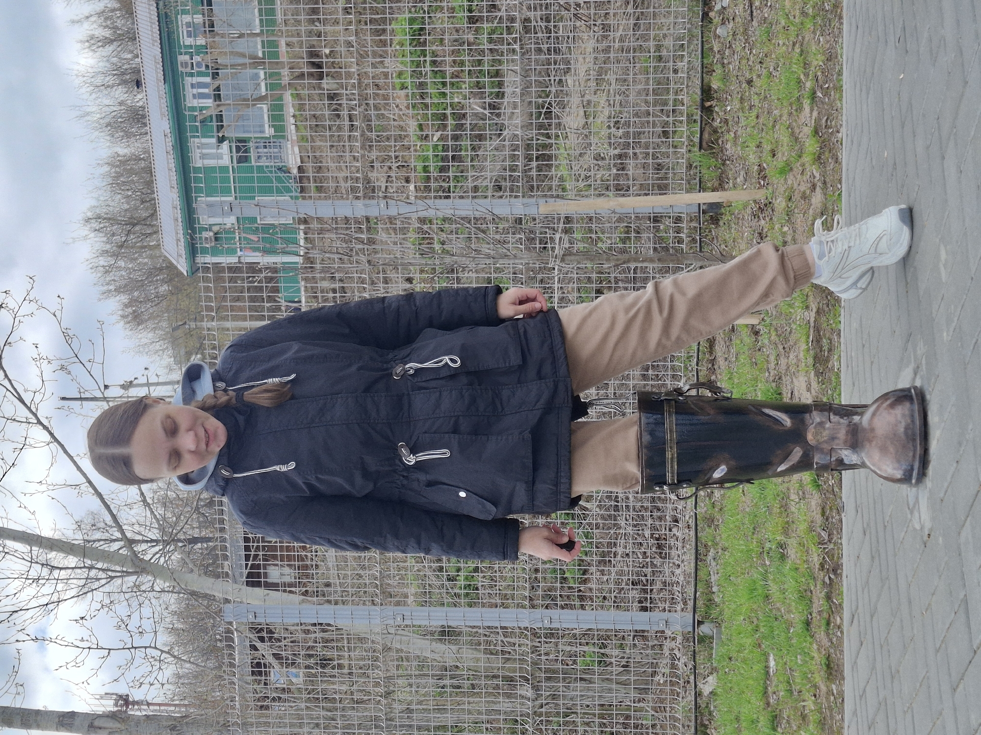 A young person stands outdoors, looking downwards with a soft smile. They have light brown hair styled in two braids. They are dressed in a dark blue hooded jacket with light blue interior trim and beige trousers. On their left foot, they wear a white athletic shoe. Their right leg is bent, and their right foot rests inside a large, dark, ornate boot-shaped decorative object, which serves as a prop or pedestal on the ground.
The background features a tall wire mesh fence. Beyond the fence, there are numerous bare trees, indicating either early spring or late autumn, interspersed with patches of green grass. In the farther background, a multi-windowed green building is visible through the sparse foliage. The ground beneath the person is paved with grey rectangular blocks. The sky above is overcast, suggesting a daytime setting with diffused light. No visible text is present in the image. The scene is located in Sarapul, Russia.