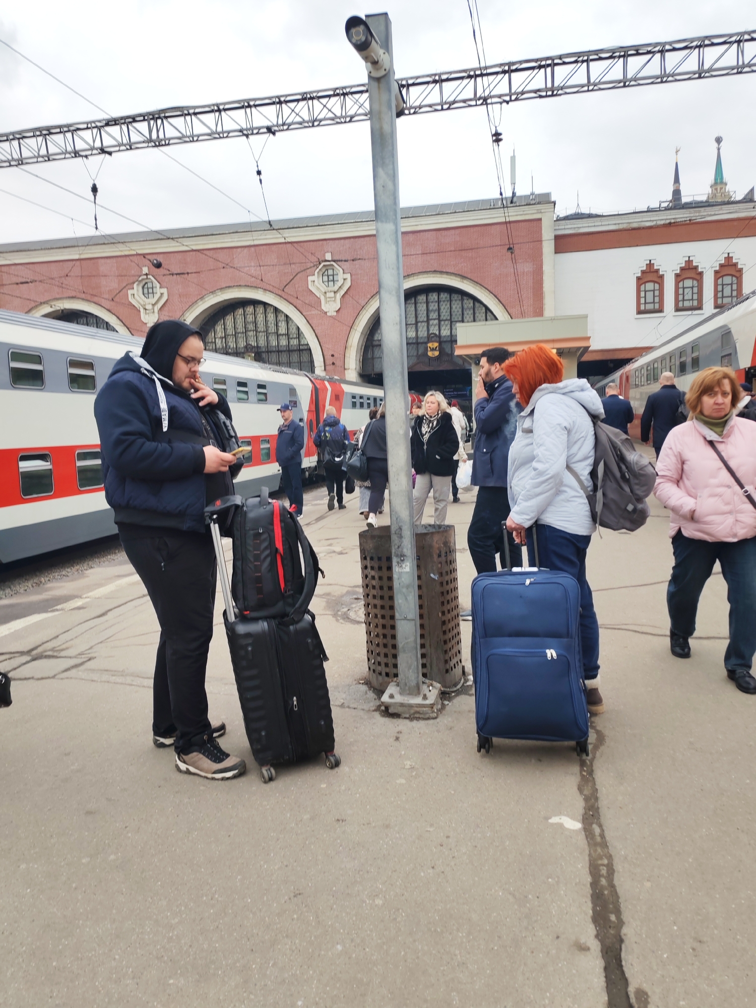 This media file depicts a bustling scene at a train station platform in Moscow, Russia, under an overcast sky. In the foreground, on the left, a man wearing a dark blue hooded jacket, glasses, and black track pants is engrossed in his smartphone, holding a lit cigarette in his mouth. He stands next to a black rolling suitcase with a black backpack attached. To the right, a woman with prominent red hair, dressed in a light grey hooded jacket and dark jeans, pulls a blue rolling suitcase and wears a grey backpack.
A tall metal pole with a security camera is centrally located, with a cylindrical metal waste bin at its base. Several other people, appearing to be travelers, populate the platform in the mid-ground and background. One man is also seen smoking behind the central pole, and a woman in a pink puffer jacket stands on the far right.
To the left, a train with white and red livery is parked at the platform. The background showcases a large, architecturally distinct station building featuring prominent red brick arches, decorative white window frames, and arched windows. A gold spire is visible on the roofline of a distant building on the right, a detail consistent with Russian architecture. Overhead power lines and a metal gantry structure span across the tracks, indicating an electrified railway system. The scene captures the typical activity of a busy train terminal.