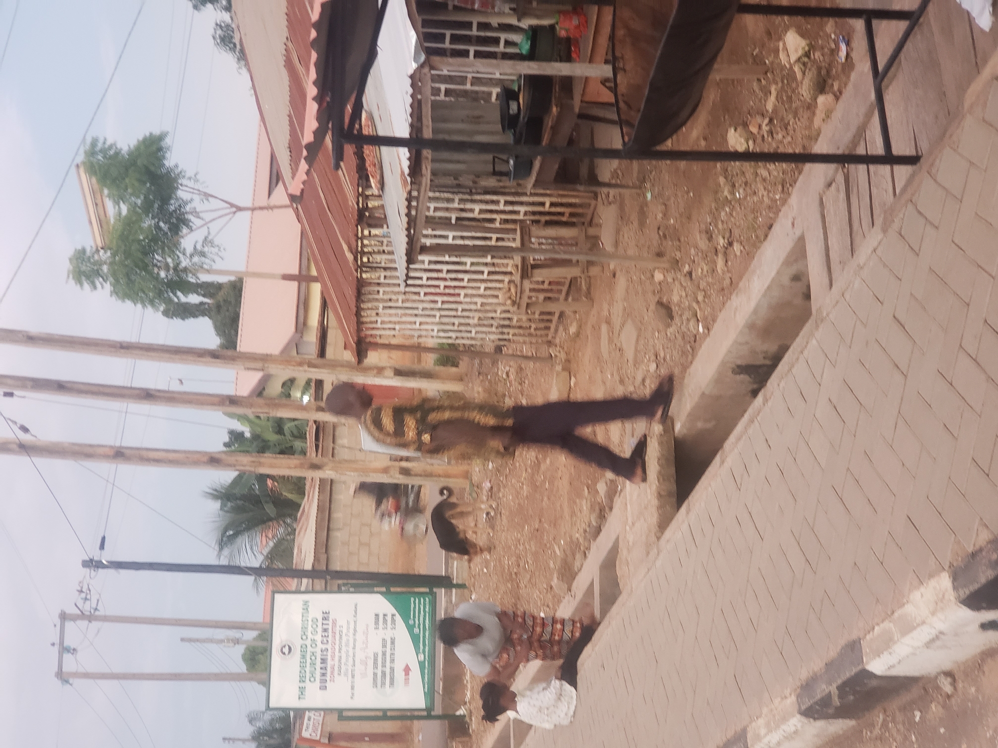 The image depicts a street scene in Narayi, Nigeria. In the foreground, a paved walkway with a concrete drainage channel runs alongside a dirt area. A young girl in a white shirt with black polka dots is sitting on the walkway, and an adult woman in a patterned skirt and light-colored top is attending to her. A man in a patterned shirt and dark pants is walking on the edge of the walkway, stepping over the drainage. A dog is visible in the background, grazing in the dirt.
Behind the people and the dog, there is a street sign for "THE REDEEMED CHRISTIAN CHURCH OF GOD DUNAMIS CENTRE ZONAL HEADQUARTERS". Further back, buildings with corrugated iron roofs are visible, as well as electrical poles and wires. Some of these buildings appear to be informal market stalls. The overall environment suggests a somewhat arid or dusty urban setting.