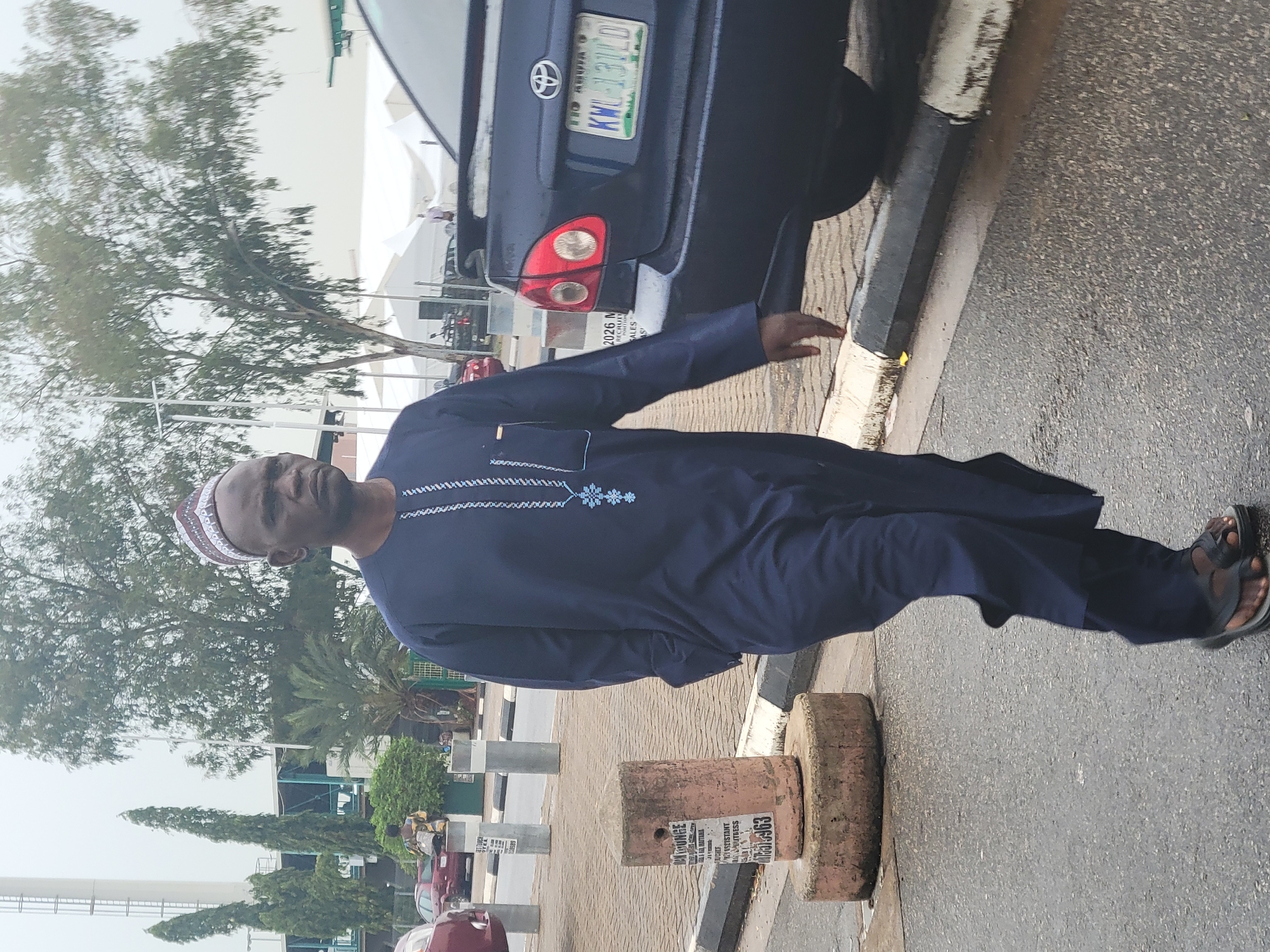 A man wearing a blue African suit and a white kufi cap stands on a city street. The license plate on the car behind him says "FCT ABUJA KNU-131LD." A cement bollard in front of him has an advertisement for "ML Lounge Bautama Assistant waitress" along with a phone number that ends in "75963." The background consists of more bollards, trees, other parked cars, and a white tent.