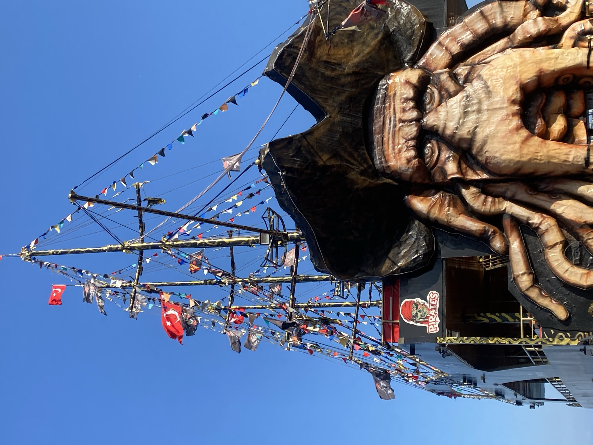 The image displays a highly decorated pirate ship under a clear blue sky, likely in Alanya, Turkey. The ship is adorned with numerous colorful triangular bunting flags strung across its masts and rigging. Several national flags of Turkey, featuring a red background with a white star and crescent, are prominently displayed among the bunting, alongside black pirate flags with skull and crossbones emblems.
A striking feature of the vessel is a colossal, intricately sculpted kraken or giant squid figure dominating the side of the ship. This sculpture is rendered in shades of brown and beige, mimicking organic textures, with visible suckers on its massive tentacles and a detailed head. The creature appears to be clinging to the ship, adding a dramatic and adventurous theme.
The ship itself has a dark hull with gold-patterned railings and details. A red banner with a white skull and crossbones emblem is visible on the ship's side, bearing the text "PIRATES" in white letters. The bright, even lighting suggests it is daytime with sunny weather. The overall scene evokes a festive, adventurous atmosphere, characteristic of a tourist attraction or themed excursion boat.