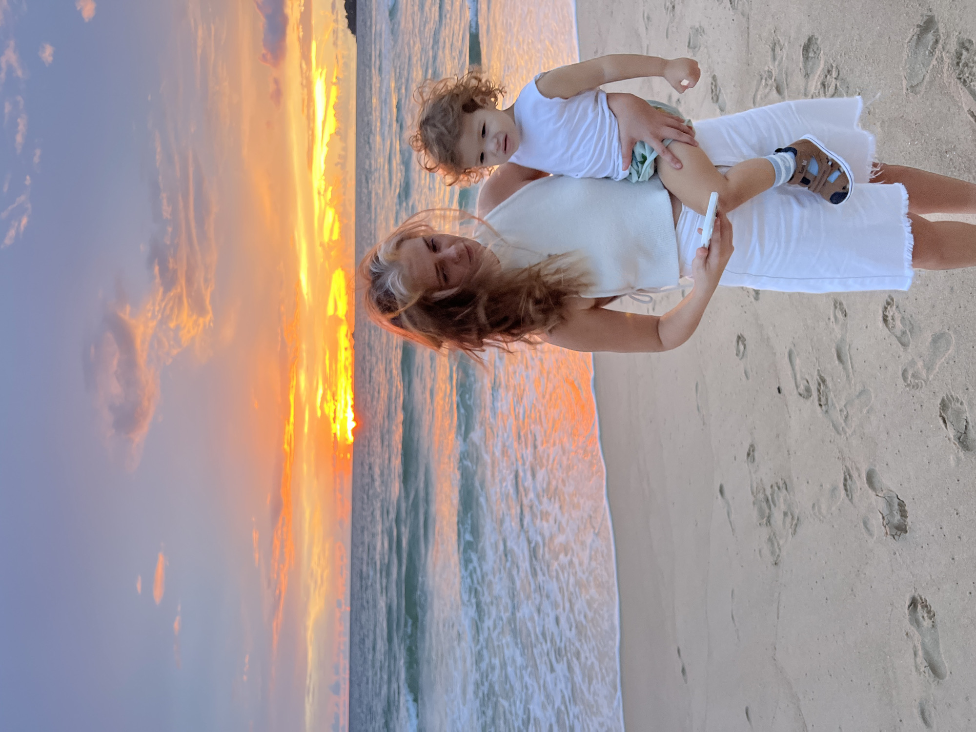 This media file depicts a mother and her child on a sandy beach during sunset. The mother is carrying the child on her shoulders, and both are dressed in white clothing. The child, with curly hair, is wearing a white tank top and light-colored shorts, while the mother has on a white top and a white skirt. The child is wearing brown sandals with blue accents and striped socks. The scene is set against a backdrop of the ocean and a colorful sky, with the sun setting and casting a warm glow. The sky is filled with clouds that are illuminated by the setting sun, creating a dramatic and beautiful display of orange, yellow, and purple hues. Gentle waves are washing ashore on the sandy beach, leaving ripples and foam. Footprints are visible in the sand, suggesting recent activity on the beach. The overall atmosphere appears peaceful and joyful, with the mother and child enjoying a moment together at the beach. The time of day is clearly sunset due to the lighting conditions and the colors in the sky. There is no visible text in the image.